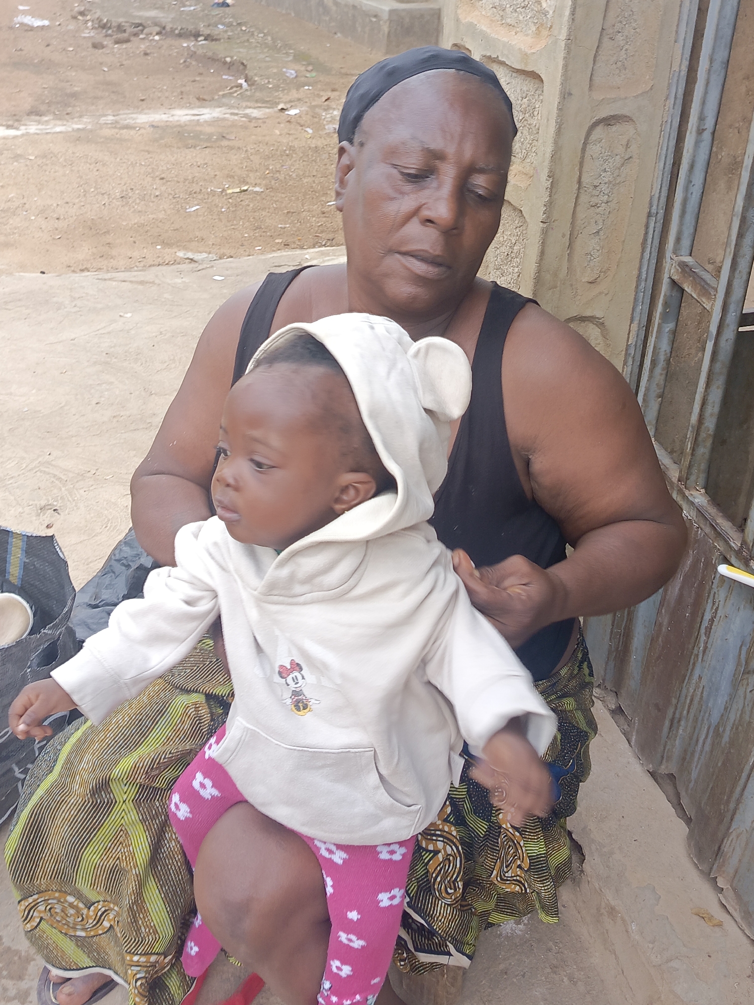 In Akwanga, Nigeria, a woman in a black tank top and patterned skirt sits outdoors, holding a baby wearing a white hooded sweatshirt and pink leggings with white patterns. The woman wears a black head covering. The background shows a dirt ground surface and a textured wall. There is a metal door or gate to the right.