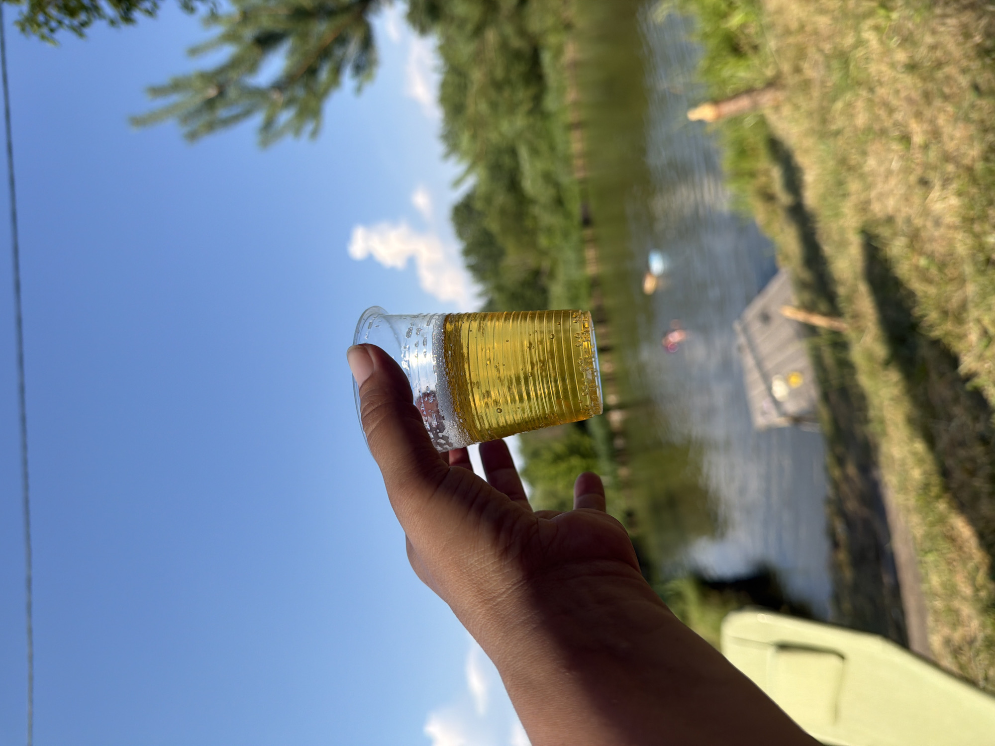 The image shows a close-up of a hand holding a clear plastic cup filled with a yellow, bubbly liquid, likely a beverage like beer or soda. The cup has ridged sides. The hand and cup are angled upwards against a bright blue sky with a few wispy clouds. In the background, out of focus, is a body of water, possibly a river or lake, lined with lush green trees. The sunlight suggests it is a sunny day, likely daytime. There are hints of a dock or wooden structure on the water, and some people are visible in the water as small, colorful figures, suggesting an outdoor recreational setting. The overall mood is relaxed and summery. The image was taken in Kamianka-Buzka, Ukraine.