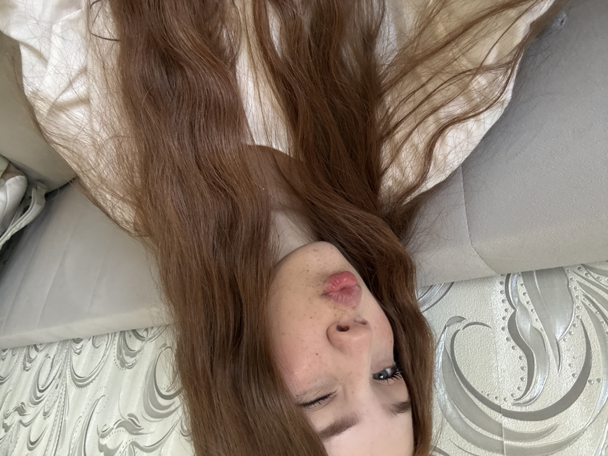 A close-up, horizontally oriented shot captures a young person with long, flowing reddish-brown hair spread across the frame. Their head is positioned towards the bottom right, with their face clearly visible. The person has noticeable freckles scattered across their nose and cheeks. They are winking with their left eye (viewer's left) while their lips are puckered into a playful "kiss" or "duck face" expression. Long eyelashes are visible, particularly on the open right eye.
The background primarily consists of the person's hair and light-colored elements. To the upper left, there's light, possibly white, fabric, which could be clothing or bedding. Further to the left and directly behind the person's head, there's a light gray textured surface, perhaps a sofa or headboard. The lower and central background features patterned wallpaper with an ornate, swirling design in silver-gray tones against a lighter, possibly off-white, base. No specific location cues for Naberezhnye Chelny, Russia, are discernible from the image. The overall mood conveyed is playful and informal. There is no visible text.