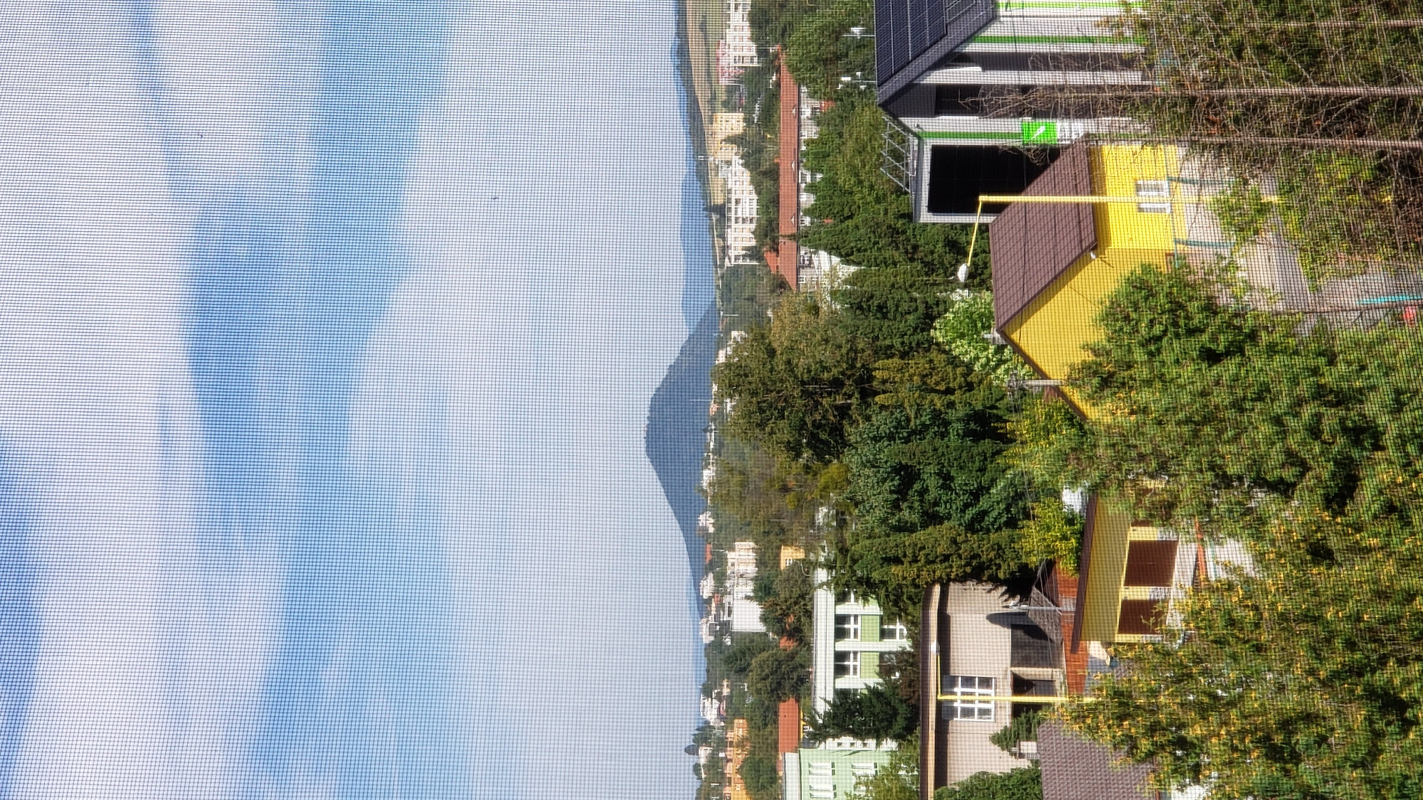 The image presents a daytime, medium-distance view of a residential area in Prešov, Slovakia, with a prominent mountain in the background. The foreground is dominated by lush green trees, some of which are in bloom with yellow flowers. Several houses are visible, including a bright yellow house with a brown roof and white windows, and a partially visible building with a dark, sloping roof and vertical green and white siding. In the middle ground, a light green, multi-story building with white window frames stands out. Further back, a cluster of apartment buildings with varied colors and window styles are nestled among more trees. The distant background features a large, dark, conical mountain under a bright blue sky dotted with white clouds. The scene is viewed through a window screen, which creates a subtle grid pattern over the entire image. There are no people or animals clearly visible in the frame.
