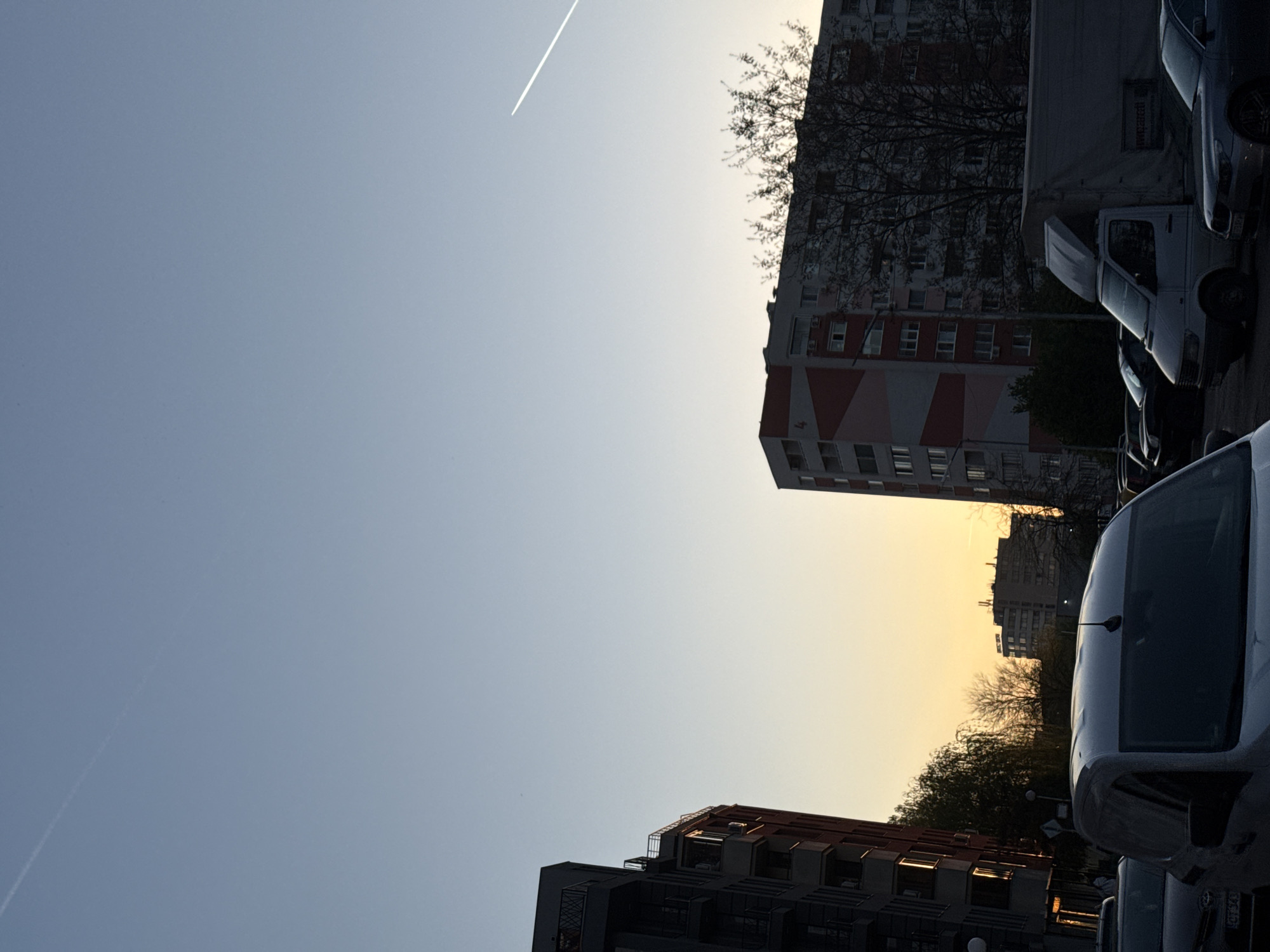 The image depicts an urban scene in Sofia, Bulgaria, at what appears to be either dawn or dusk, based on the gradient color of the sky. The view is primarily upwards, showing several apartment buildings of varying heights and architectural styles. One building has a distinct red and white striped pattern. There is evidence of trees scattered among the buildings, some with bare branches. Parked white vans are visible in the foreground. A contrail streaks across the upper part of the sky.