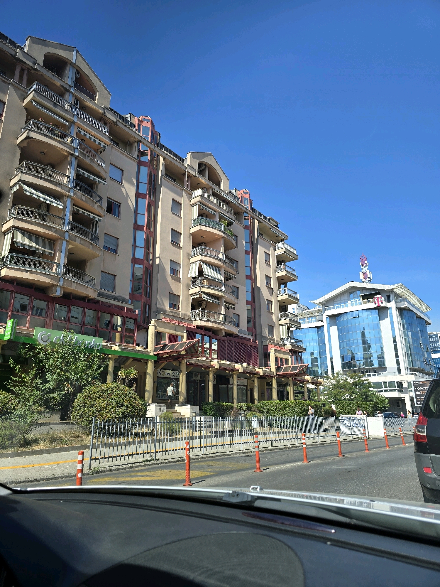 This is a medium-angle, eye-level, outdoor shot of a city street in Podgorica, Montenegro, on a sunny day. In the foreground, the dashboard of a car is visible, indicating the photo was taken from within a vehicle. The street itself is paved with asphalt and marked with yellow lines. A line of orange traffic cones and a metal fence separate the road from a sidewalk and building fronts.
The primary subjects are two multi-story buildings. The building on the left is a beige, apartment-style building with multiple balconies, each equipped with striped awnings. The ground floor of this building features a green awning with the logo "NLB Banka" clearly visible, along with storefronts. Dense green foliage is present in front of this building.
The building on the right is a modern, glass-fronted office building with a distinctive white facade and a prominent antenna topped with red lights. The glass reflects the clear blue sky. Several people are visible on the sidewalk in front of the buildings, appearing to be walking or standing. A dark-colored car is parked on the right side of the street, partially visible in the frame. The overall impression is one of urban commercial and residential activity under a bright, clear sky.