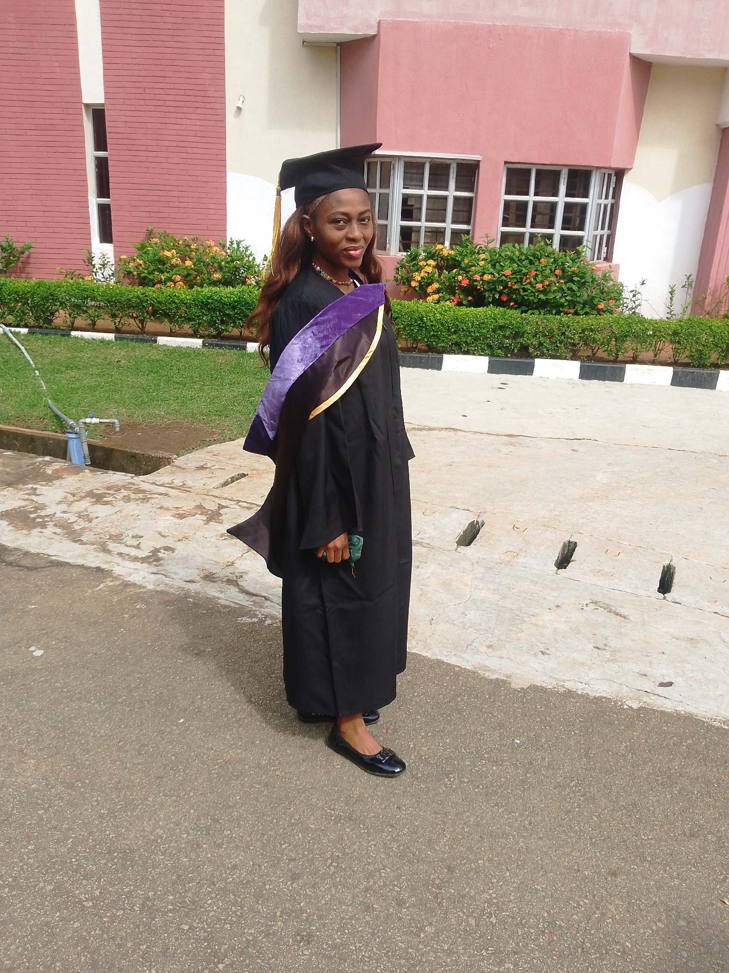 This media file captures a young woman dressed in a black graduation gown and cap, complete with a purple and gold sash. She stands on an outdoor paved area, likely on a university campus, given the background. Behind her, a building with pink and beige walls and large, multi-paned windows is visible. A neat row of green bushes with vibrant yellow and orange flowers separates the paved area from the building. A patch of green grass is to the left of the frame, with a grey hose and a blue water tap extending from it. The lighting suggests it is daytime, and the weather appears fair. The woman is looking towards the camera with a slight smile, possibly expressing pride or happiness on her graduation day. The scene is set in Agbani, Nigeria.