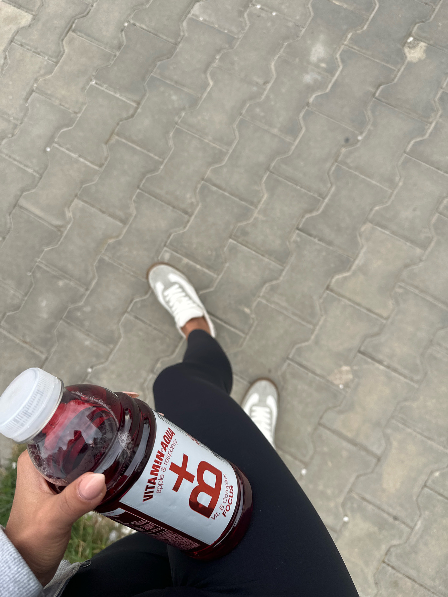 A first-person perspective shot shows a hand holding a bottle of "VITAMIN AQUA" flavored apple and raspberry drink. The bottle has a red liquid and a white cap. The person is wearing black leggings and white sneakers, suggesting they are engaged in physical activity. The background consists of interlocking concrete paving stones, commonly found in urban environments. The scene appears to be on a sidewalk in Sofia, Bulgaria, given the typical style of paving and the presence of an urban setting.