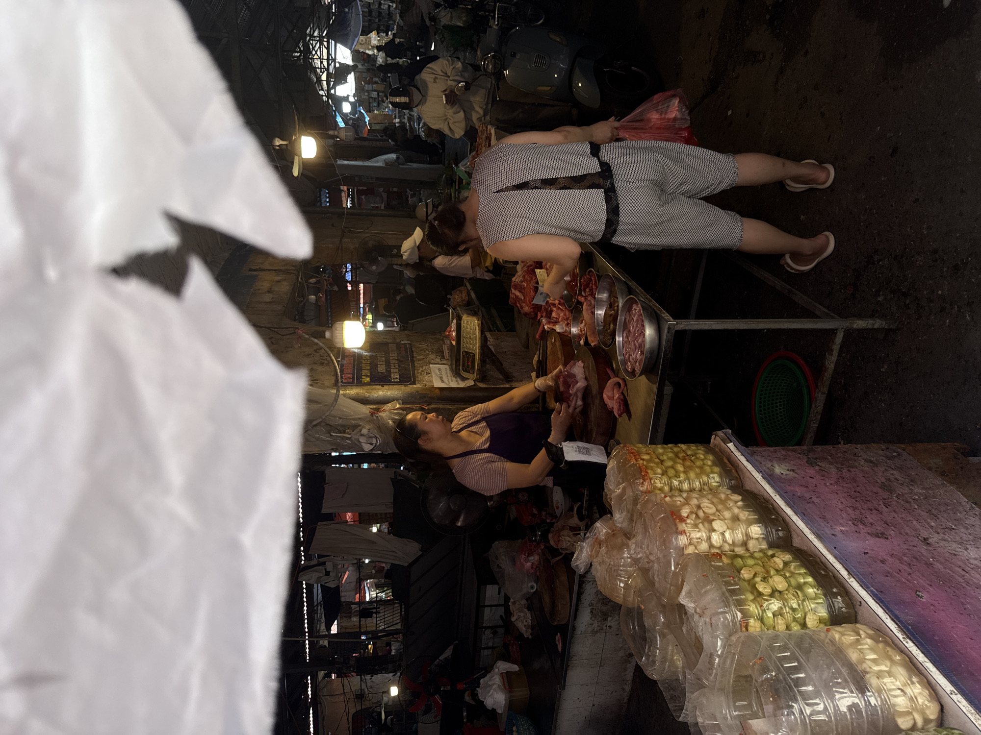 The image depicts a bustling street scene in Hà Nội, Vietnam. Two women are prominently featured: one, wearing a striped jumpsuit, appears to be a customer examining raw meat at a vendor's stall, while the other, dressed in a purple top, is likely the vendor, actively handling and preparing the meat. The stall itself is well-stocked with an array of goods, including vegetables packaged in plastic containers. The surroundings indicate a densely populated urban environment, with motorbikes parked in the background and various shops and stalls lining the street. The overall atmosphere suggests a vibrant, active commercial area typical of a busy Vietnamese city.