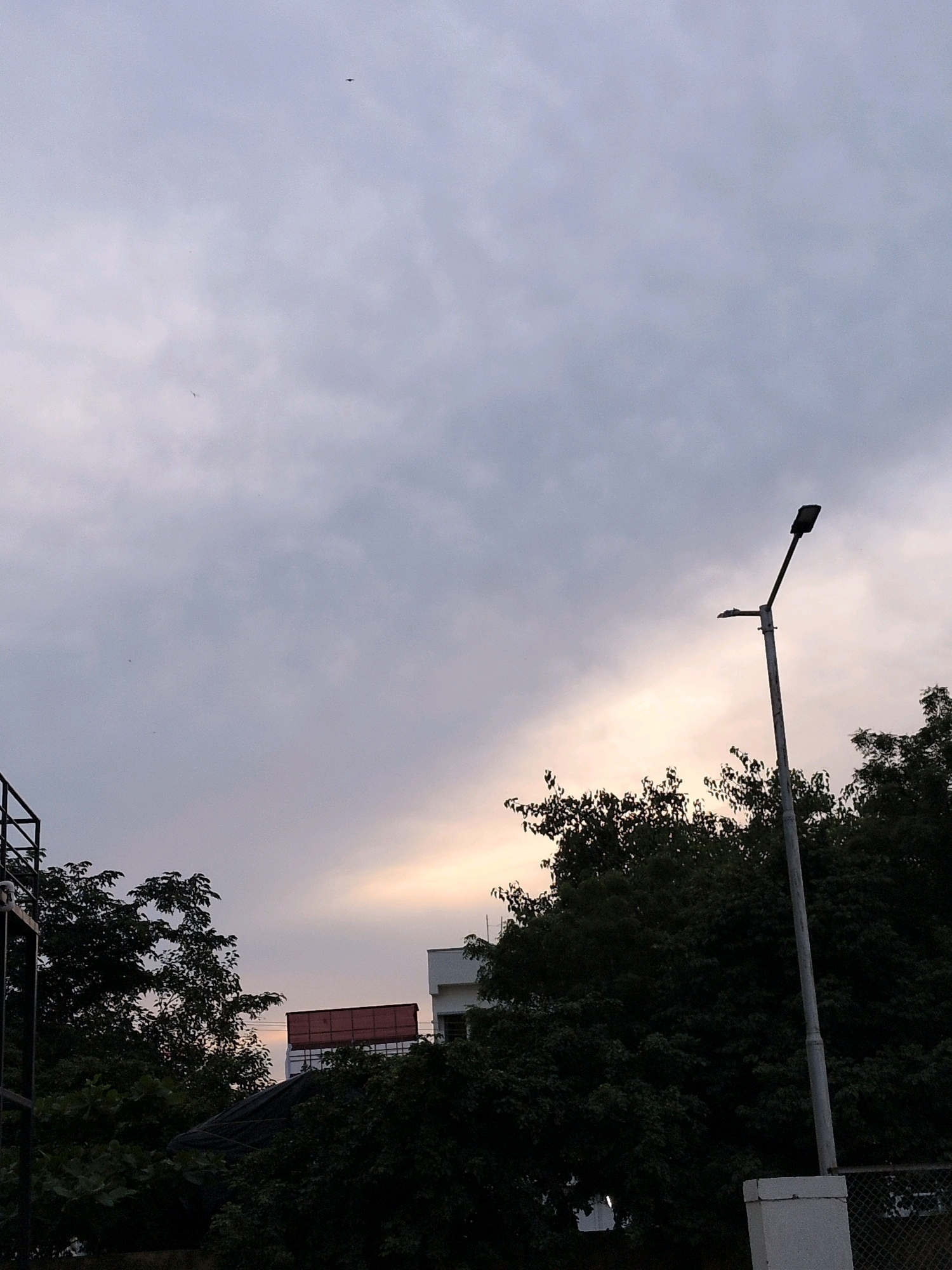 A street lamp stands against a cloudy sky in Abdullapurmet mandal, India. The lamp is a tall, grey pole with a black light fixture at the top, and a smaller arm extends from the pole below the light. The sky is filled with various shades of grey and white clouds, with a soft glow of light, possibly from the setting sun, visible in the upper right portion of the frame. Dense, dark green trees fill the bottom and right side of the image, obscuring some of the lower structures. To the left, part of a black metal structure, possibly a railing or part of a building, is visible. In the background, a red-topped structure, likely a container or part of a building, sits atop a white vehicle. Another building with a white facade is visible behind the trees on the right. A few small, dark specks, possibly birds or insects, are visible in the sky.