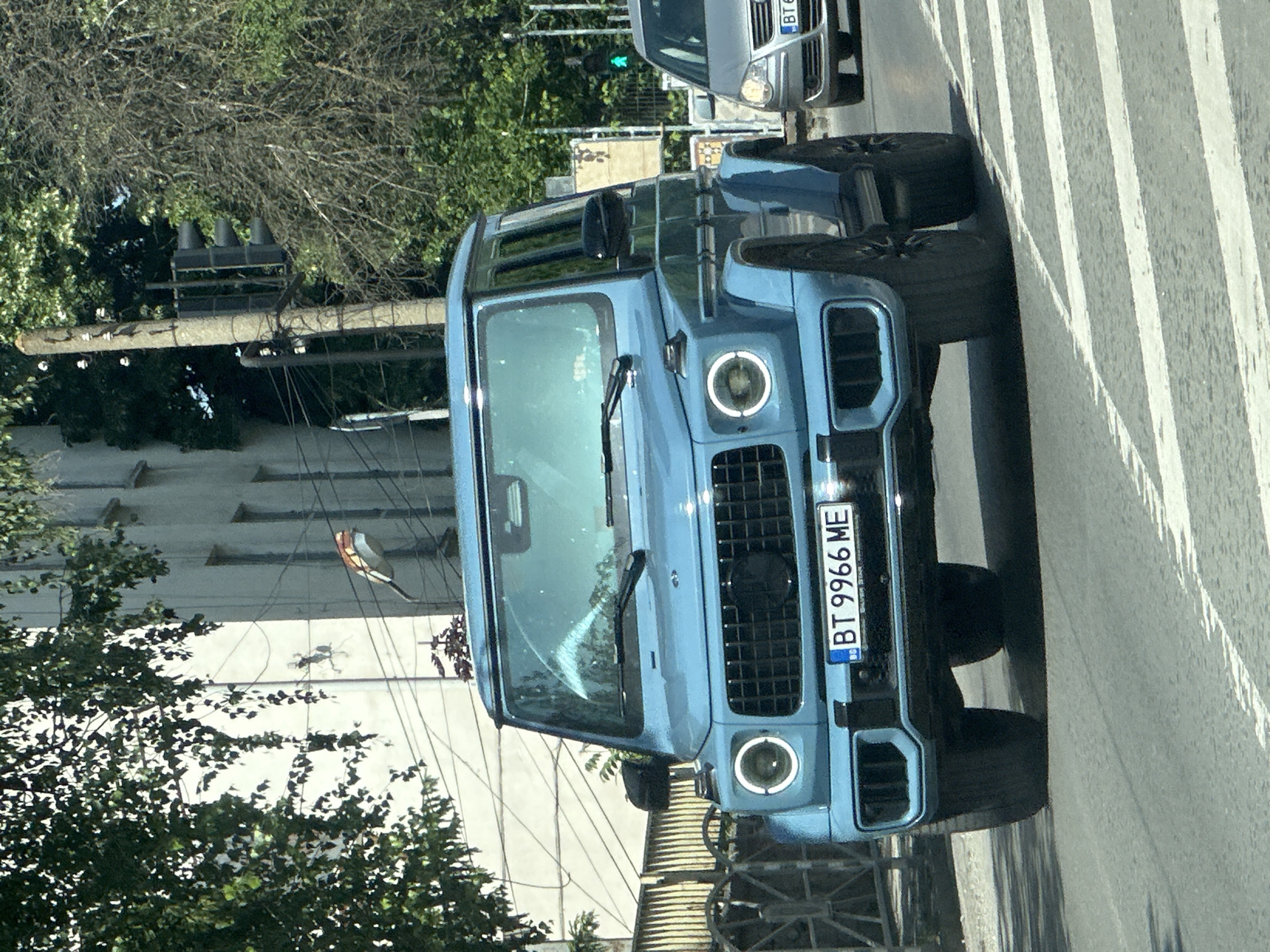 A blue Mercedes-Benz G-Class is parked on a crosswalk in Veliko Tarnovo, Bulgaria. The car's license plate reads "BT 9966 ME." A grey Volkswagen Touran is parked ahead of it. Traffic lights with a green light are visible in the background. A utility pole and overhead wires are on the left side of the image, near a building with several windows. Trees are visible in the foreground.