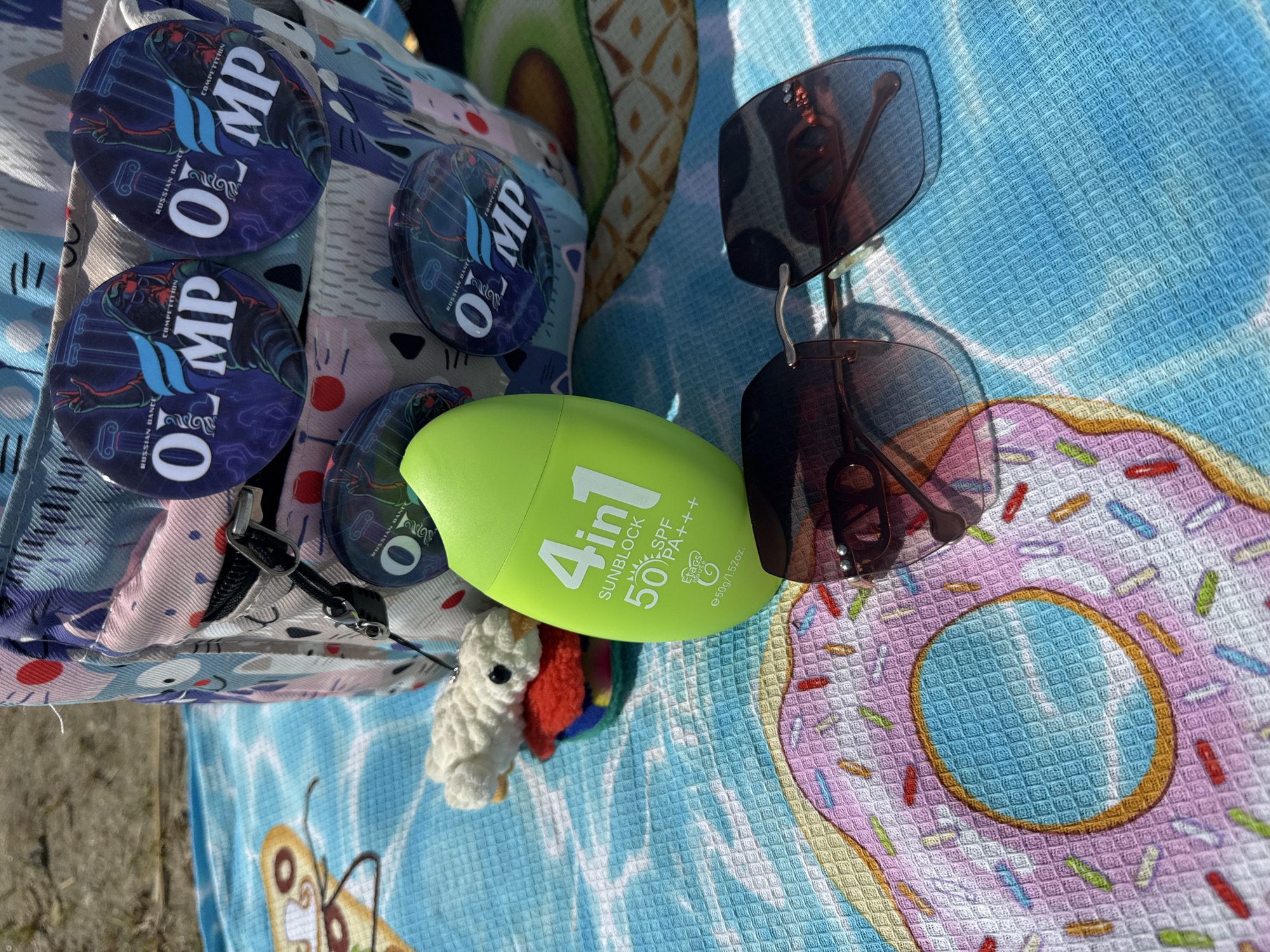 The image depicts a close-up, outdoor scene with a patterned beach towel as the primary surface. On the towel, a bright green bottle of "4 in 1 Sunblock 50 SPF PA+++" is prominently displayed. Next to the sunblock, a pair of modern sunglasses with dark lenses rests. In the background, a colorful backpack adorned with circular pins featuring the text "RUSSIAN DANCE COMPETITION OLIMP" is visible. Also on the towel, a small, fluffy white object resembling a bird or a stuffed toy is situated near the sunblock. The beach towel itself has a vibrant design of a pink frosted donut with colorful sprinkles, set against a blue background with a wave-like pattern. The overall environment suggests a sunny day, likely at a beach or park. There is no discernible information that places this scene specifically within the city of Chelyabinsk, Russia.