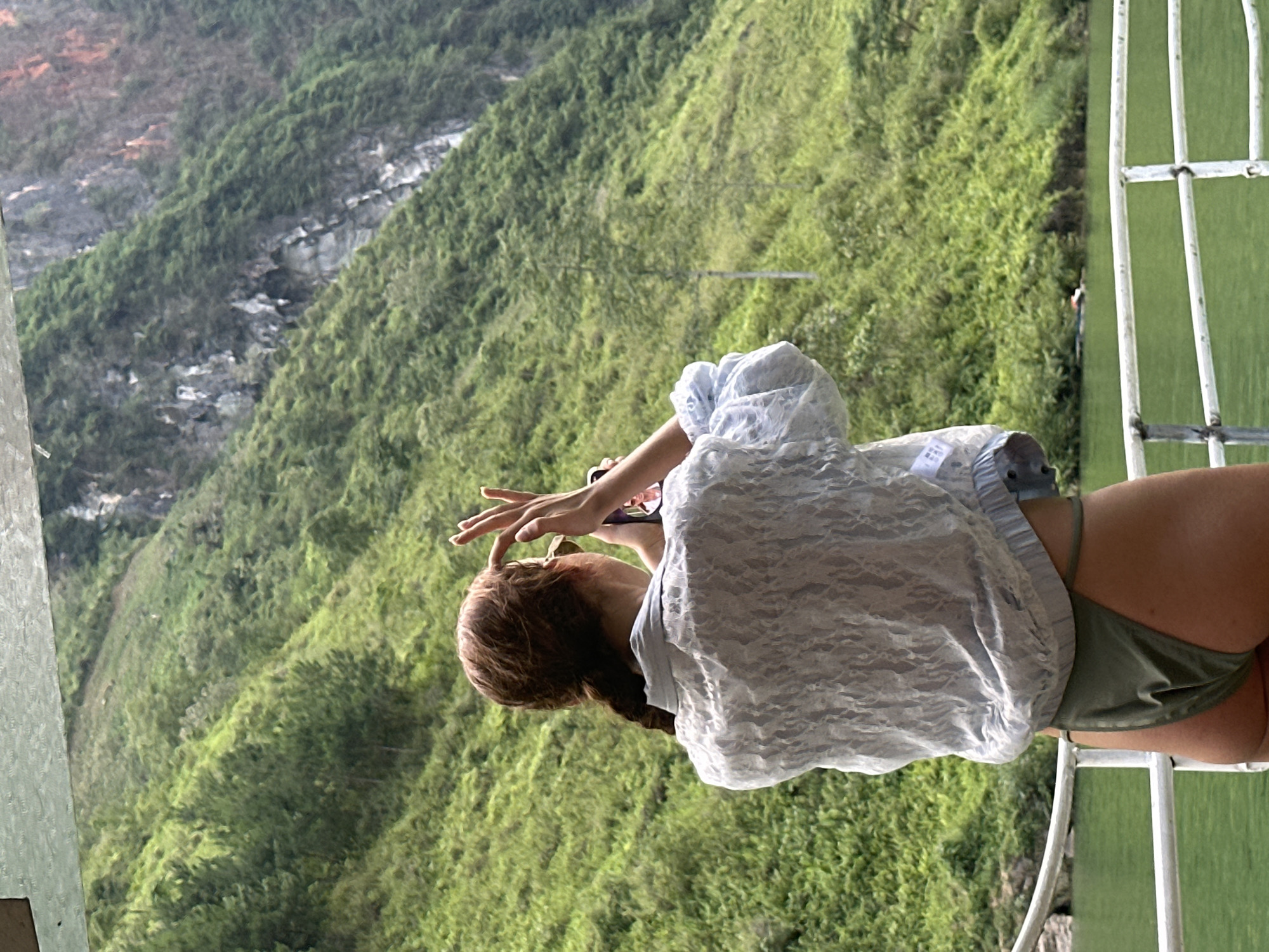 A woman with reddish-brown hair in a braid stands on a white metal structure in Đồng Văn Commune, Vietnam. She wears a light blue, lace-like top over a dark green bathing suit bottom. She appears to be using a smartphone to take a photo. The background is a steep, green, mountainous landscape with rocky sections.