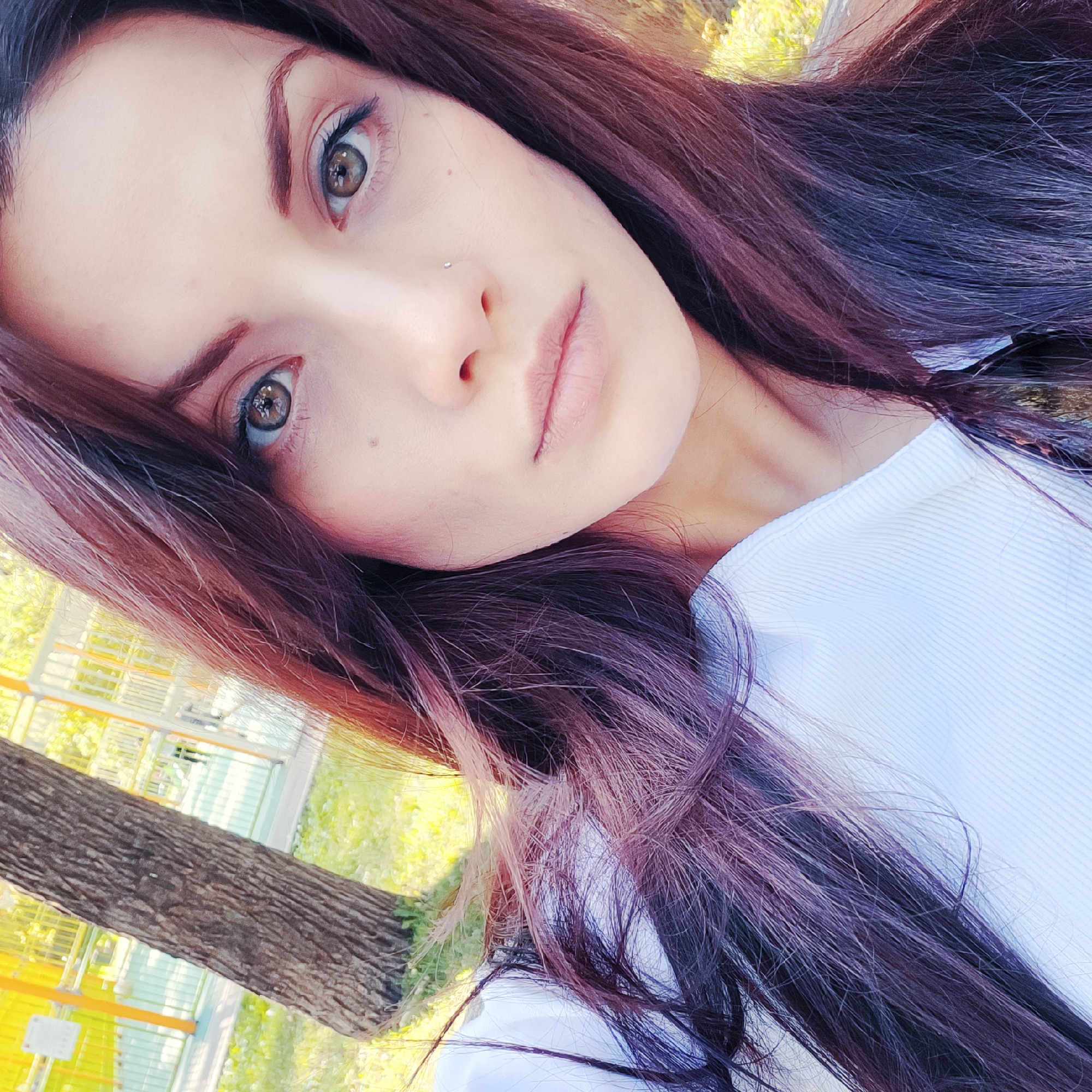 A close-up, front-facing selfie of a young adult female with long, dark auburn hair. Her eyes are a greenish-brown, and she has a nose ring. She is wearing a white, ribbed top. The background is blurred but appears to be an outdoor park setting with a tree trunk on the left and a yellow structure and greenery on the right. The lighting suggests it is daytime.