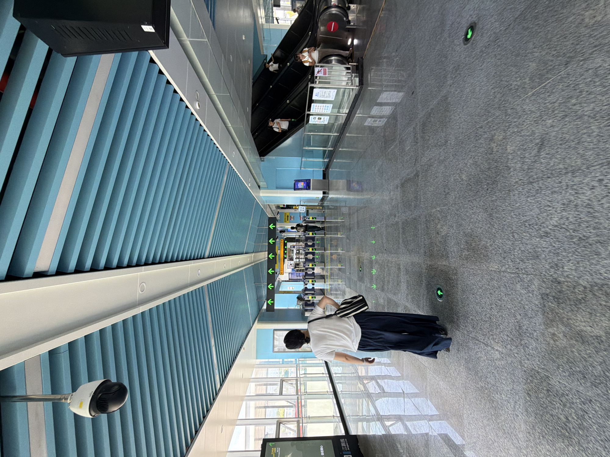 The image shows a person from behind walking down a concourse in what appears to be a modern transit station. The concourse features a light blue, slatted ceiling, a polished gray stone floor, and large windows on the left side looking out onto an exterior area. Green directional arrows are embedded in the floor, guiding pedestrian traffic. On the right side of the concourse, an escalator is visible with several people using it, as well as a large black speaker mounted to the ceiling. A security camera is also visible hanging from the ceiling near the entrance of the concourse. The overall impression is of a clean, orderly, and well-lit public space. This scene is located in the Jiangjin district of Chongqing, China, within the Jiangjin Long Line metro station.