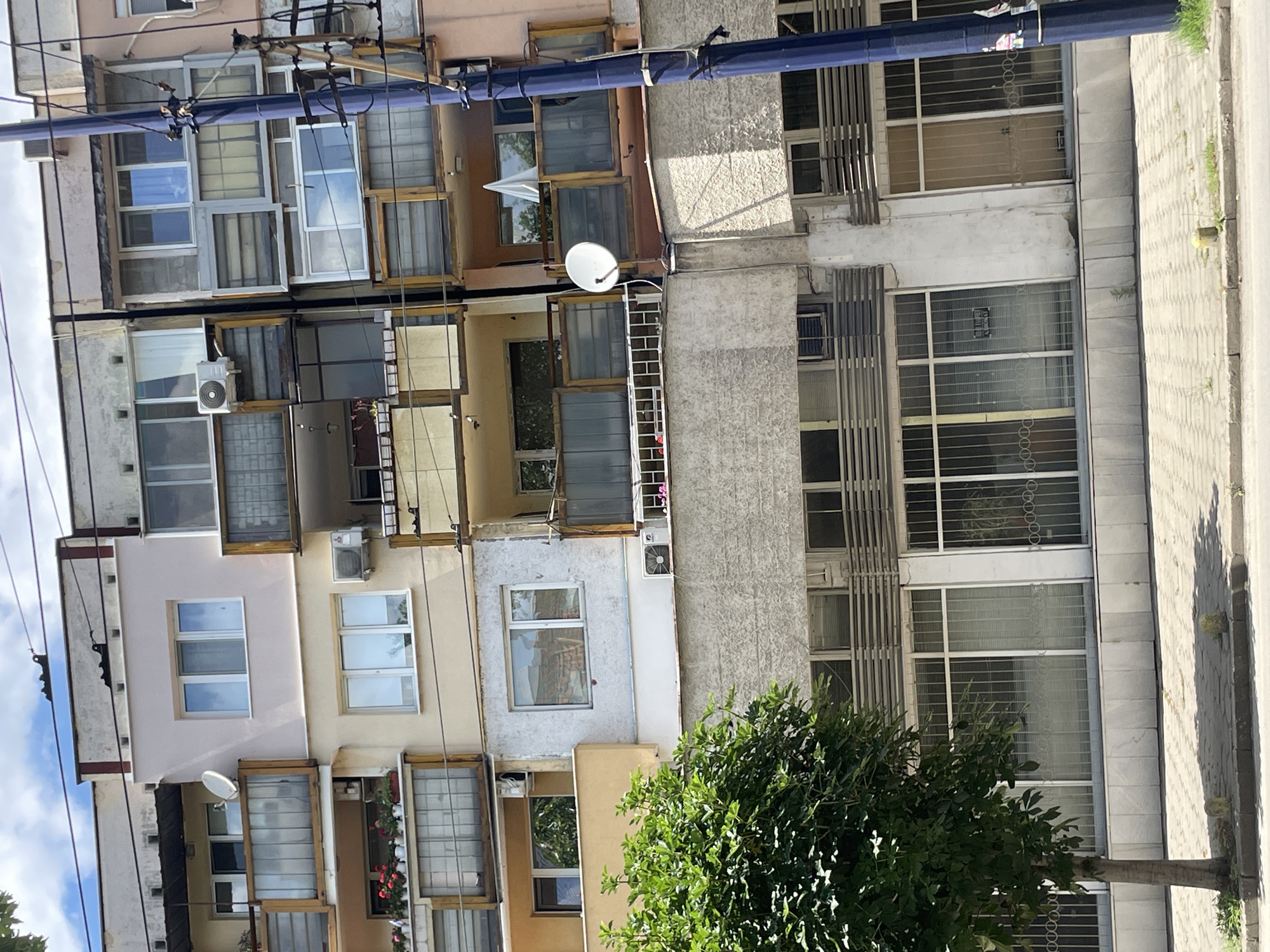 The media file depicts an outdoor scene of a multi-story building with a complex facade, characterized by numerous windows, balconies, and varying architectural styles. The building appears to be an apartment complex or a mixed-use structure, with many windows indicative of individual units. Some windows are open, revealing interior spaces and reflections of the sky. An air conditioning unit is visible mounted on one of the walls. Utility poles and wires run across the upper portion of the image, suggesting an urban or suburban environment.
In the foreground, the lower section of the building features balconies adorned with plants, including flowers, adding a touch of greenery. A tree with dense foliage is positioned to the right, partially obscuring some of the lower windows and the base of the building. The ground in front of the building is paved with interlocking blocks, and a curb lines the edge of what appears to be a street or sidewalk.
The scene is bathed in daylight, suggesting it was taken during the daytime. The sky visible in some windows is a light blue with some clouds, indicating fair weather. There are no people actively visible in the scene. The overall impression is a somewhat aged and perhaps densely populated residential building, with a mix of modern and older architectural elements. Some windows have metal security bars or grilles. The image is oriented vertically, and the building's facade dominates the frame. No visible text is clearly discernible.