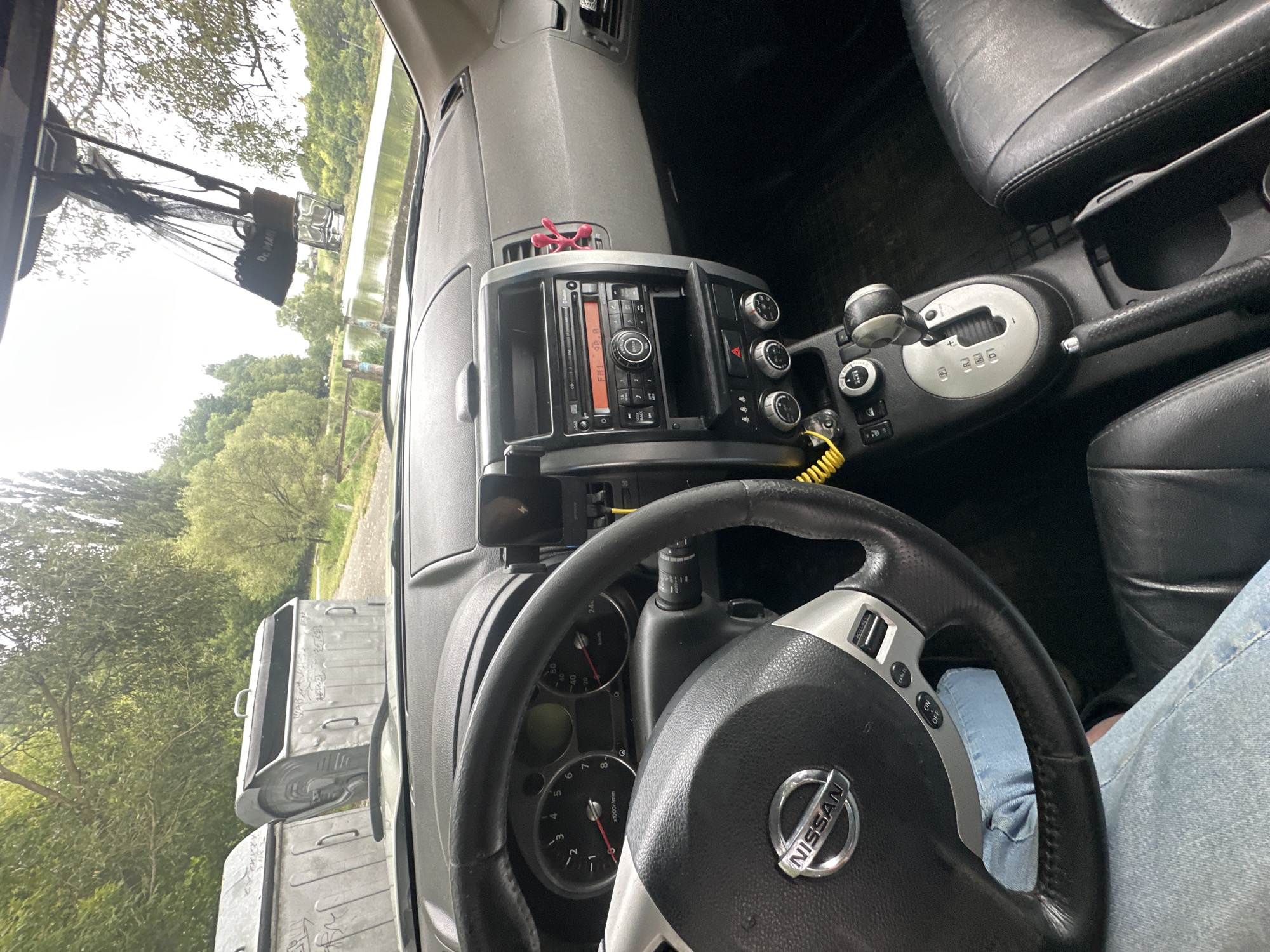 The image provides an interior view of a Nissan car, focusing on the dashboard and driver's seat. The steering wheel, with the Nissan logo, is prominent on the left. The center console features a car stereo, climate controls, and a gear shifter. A yellow cable is visible near the gear shifter. Outside the car, through the windshield, two large grey dumpsters are partially visible, situated in an outdoor environment with trees and a body of water in the background. The scene suggests the car is parked outdoors, possibly near a rural or semi-rural area. The location is identified as Makiv, Ukraine.