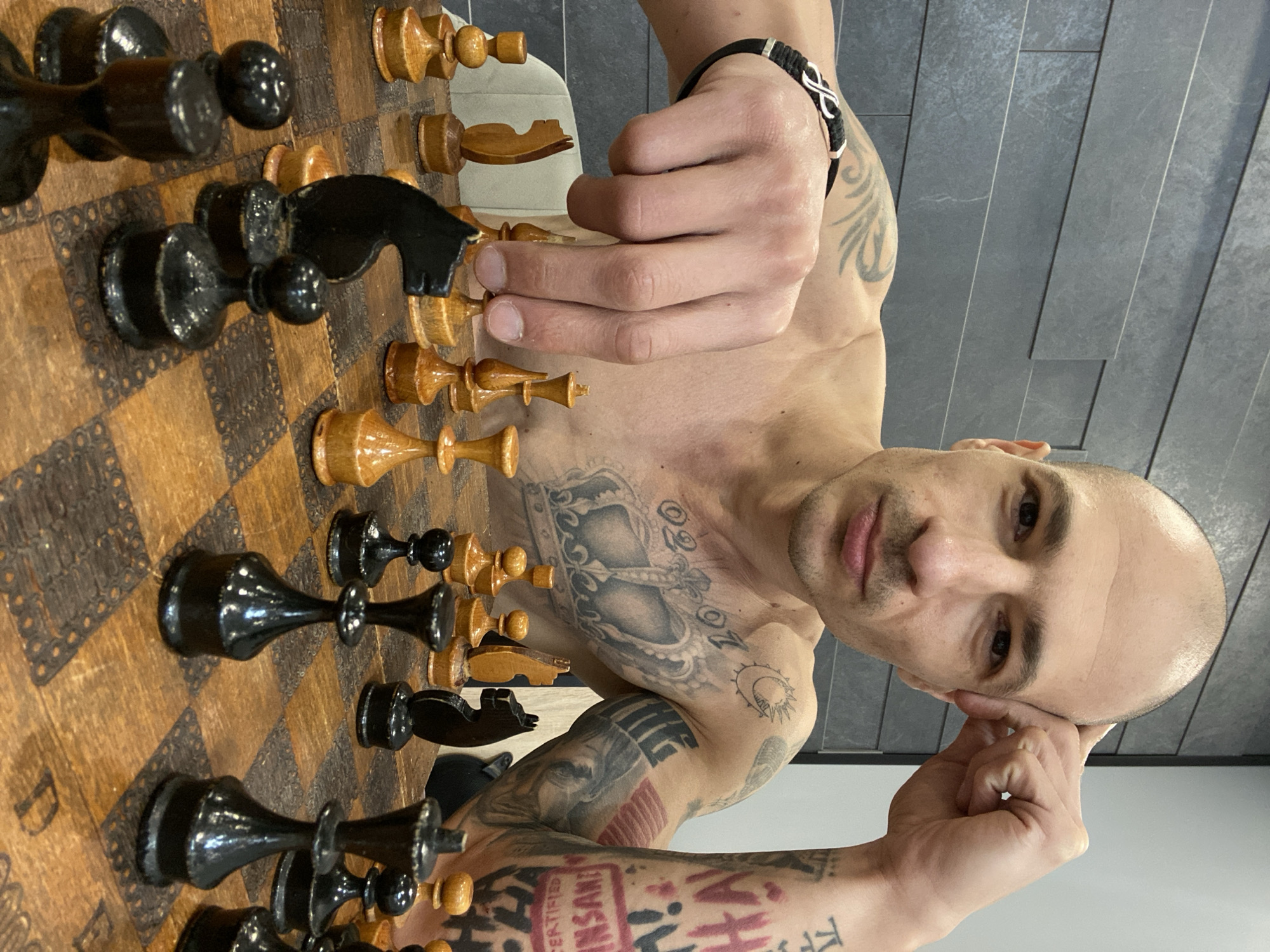 A man with a shaved head and numerous tattoos is playing chess. He is shirtless and appears to be deep in thought, with one hand resting on his head. The chess pieces are arranged on a wooden board, with black pieces on one side and light brown pieces on the other. The background is a modern wall with gray panels. The location is Pleven, Bulgaria.