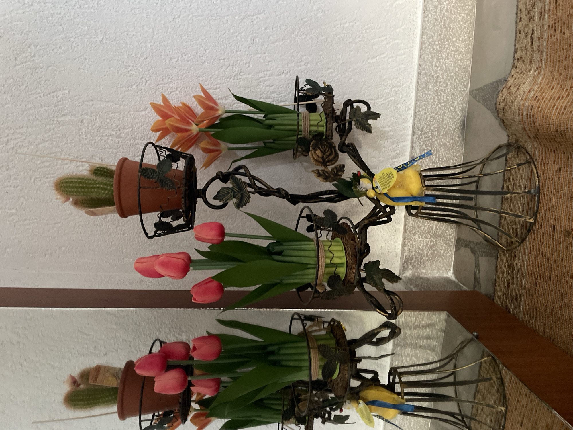 The image shows a decorative metal candelabra holding potted plants. On the top tier, there are two potted cacti. Below them, on the next two tiers, are clusters of artificial tulips, some red and some orange. The candelabra is placed indoors, near a wall with a textured white finish and a floor covered with a woven rug. A mirror is positioned to the left of the candelabra, reflecting the scene. Small yellow bird ornaments with blue ribbons are attached to the candelabra. The overall scene is domestic and decorative. The location in Zlatograd, Bulgaria, is inferred from the user's request.