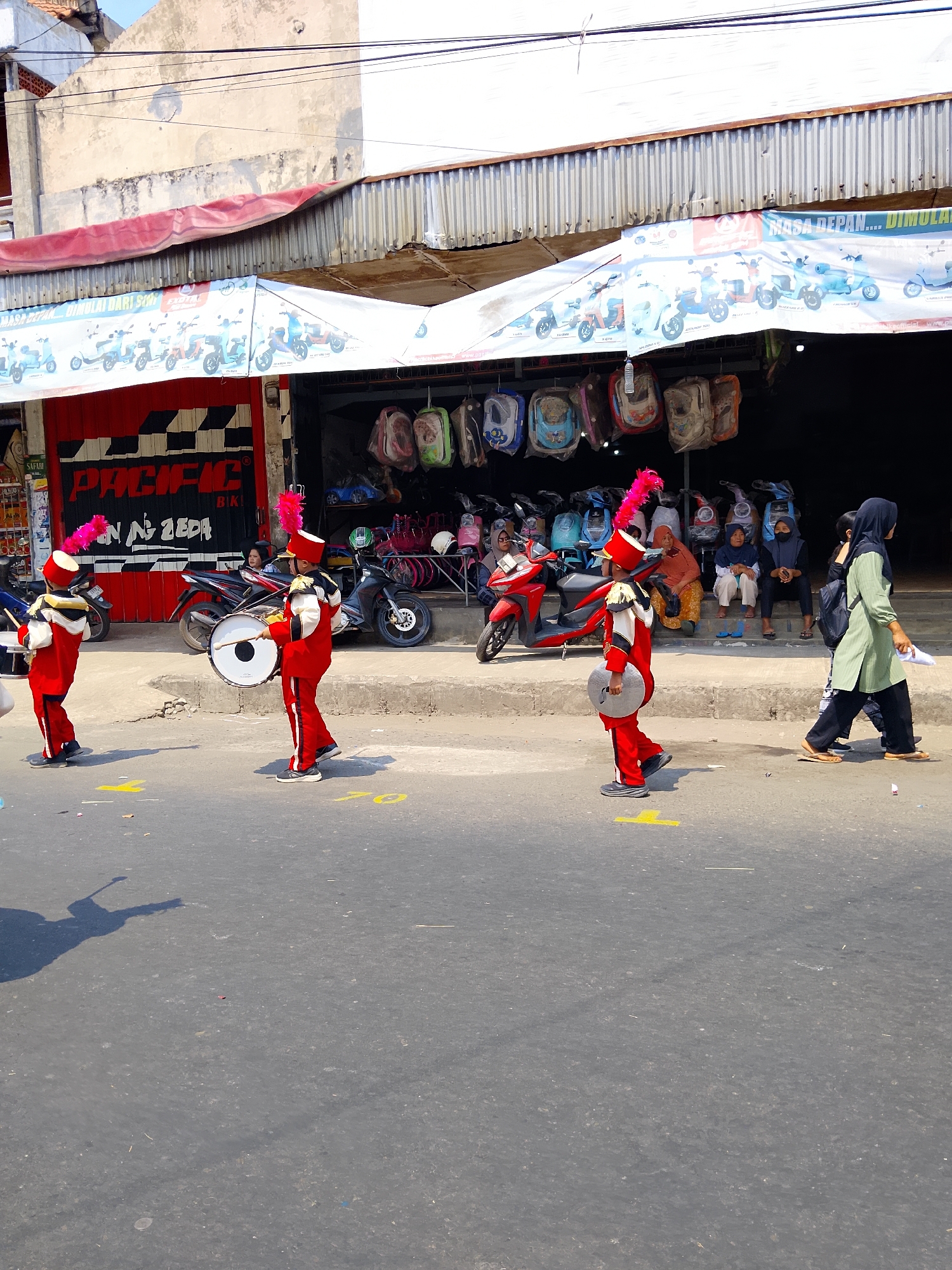 In Kertosono, Indonesia, a street scene unfolds under a bright sky. Three young individuals, dressed in vibrant red marching band uniforms with pink plumes on their hats, are actively participating in a parade. The person on the left is holding a drum, while the other two are holding cymbals. They are walking on an asphalt road, marked with yellow lines. In the background, a row of shops displays various items, including scooters and hanging bags. Several people are seated on the steps of the shops, observing the parade. A woman in a green hijab and black pants walks past in the foreground, carrying a white sheet of paper. The overall atmosphere is lively and festive.