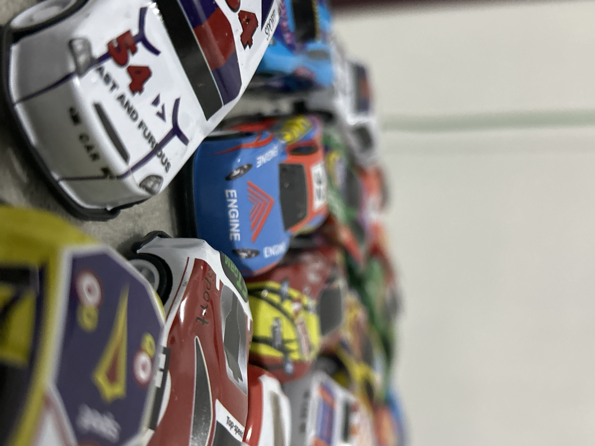 The image depicts numerous small-scale toy race cars arranged densely on a light-colored surface within an unspecified indoor location in the city of Lhokseumawe, Indonesia. In the foreground, a white toy car features the red number '54' and text 'FAST AND FURIOUS' on its hood, along with 'M CAR' below a central grill and purple stripe decals. Adjacent to it, a blue toy car is marked with 'ENGINE' in red text and has a red triangular graphic on its hood, with the number '45' visible on its roof. Further into the midground, a red toy car displays yellow and black livery, while another red and white car has 'Sport' text on its side and 'Top-speed' on its roof. Other toy cars of various colors including yellow, green, and silver are visible in the background, generally out of focus, creating a perception of depth. The background consists of a uniformly light-colored wall or vertical surface, with a faint vertical line on the left. No individuals are present in the scene.