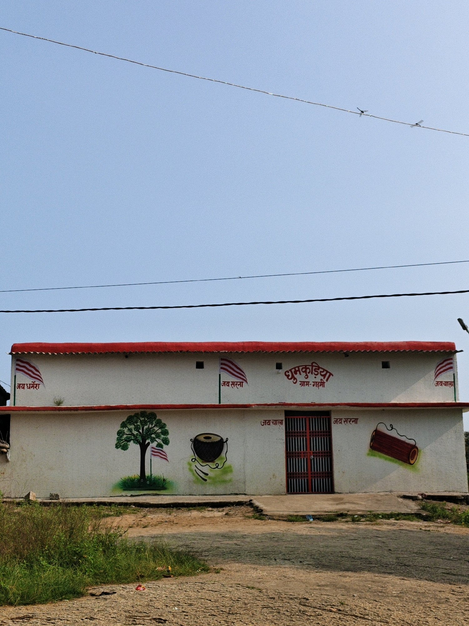 The image shows the exterior of a white building with a red roof, located in the village of Garmi, India. The building is adorned with painted murals. On the left side, a flag with red and white stripes and the Hindi text "जय धर्मेश" (Jai Dharmesh) is painted. In the center, the Hindi text "धमकुड़िया ग्राम-गढ़मी" (Dhamkudiya Gram-Garmi) is written above a red-doored entrance. To the left of the entrance is a mural of a tree with a flag at its base, and to the right is a drawing of a drum with sticks. On the right side of the building, another flag with red and white stripes and the Hindi text "जय सarna" (Jai Sarana) is painted, followed by a mural of a log. The building is surrounded by dry vegetation and a paved surface. The sky is clear and blue.