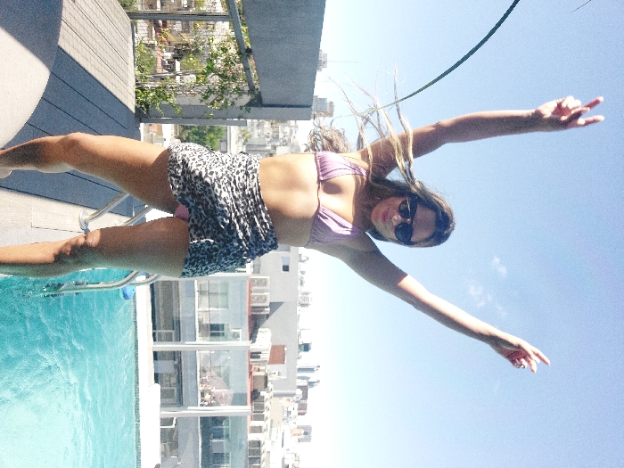A female subject stands centrally on a rooftop in Buenos Aires, Argentina, under a clear blue sky. She faces the camera, both arms raised above her head with hands forming "V" gestures. Her long, light brown hair is windswept, and she wears dark sunglasses, a light purple bikini top, and a black-and-white leopard print sarong draped over a pink bikini bottom. A slight smile is visible on her face. To her right, a light-colored composite deck extends, bordering a turquoise swimming pool with a partially submerged metal ladder visible at its edge. The background features a dense urban panorama of predominantly light-colored buildings, indicating a high vantage point within the city under bright daylight.