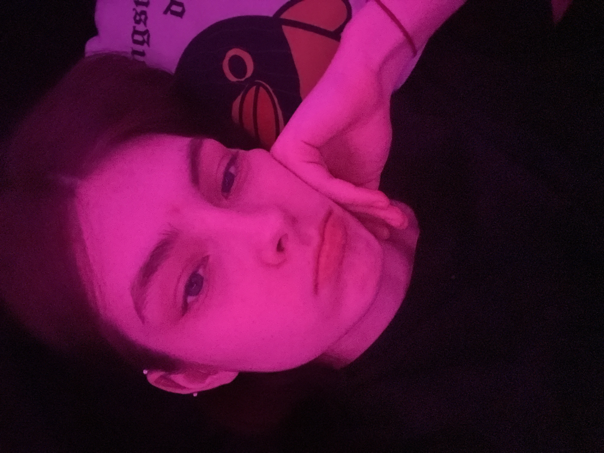 A person with light skin is positioned in a dark environment, illuminated by a pink light source. The person's face is turned towards the camera with a slightly pensive expression. Their hand is raised, supporting their face. Adjacent to the person's head is a dark-colored plush toy with a light-colored design. The person is wearing a dark-colored top. The image provides no information about the city of Saint Petersburg or any specific location.