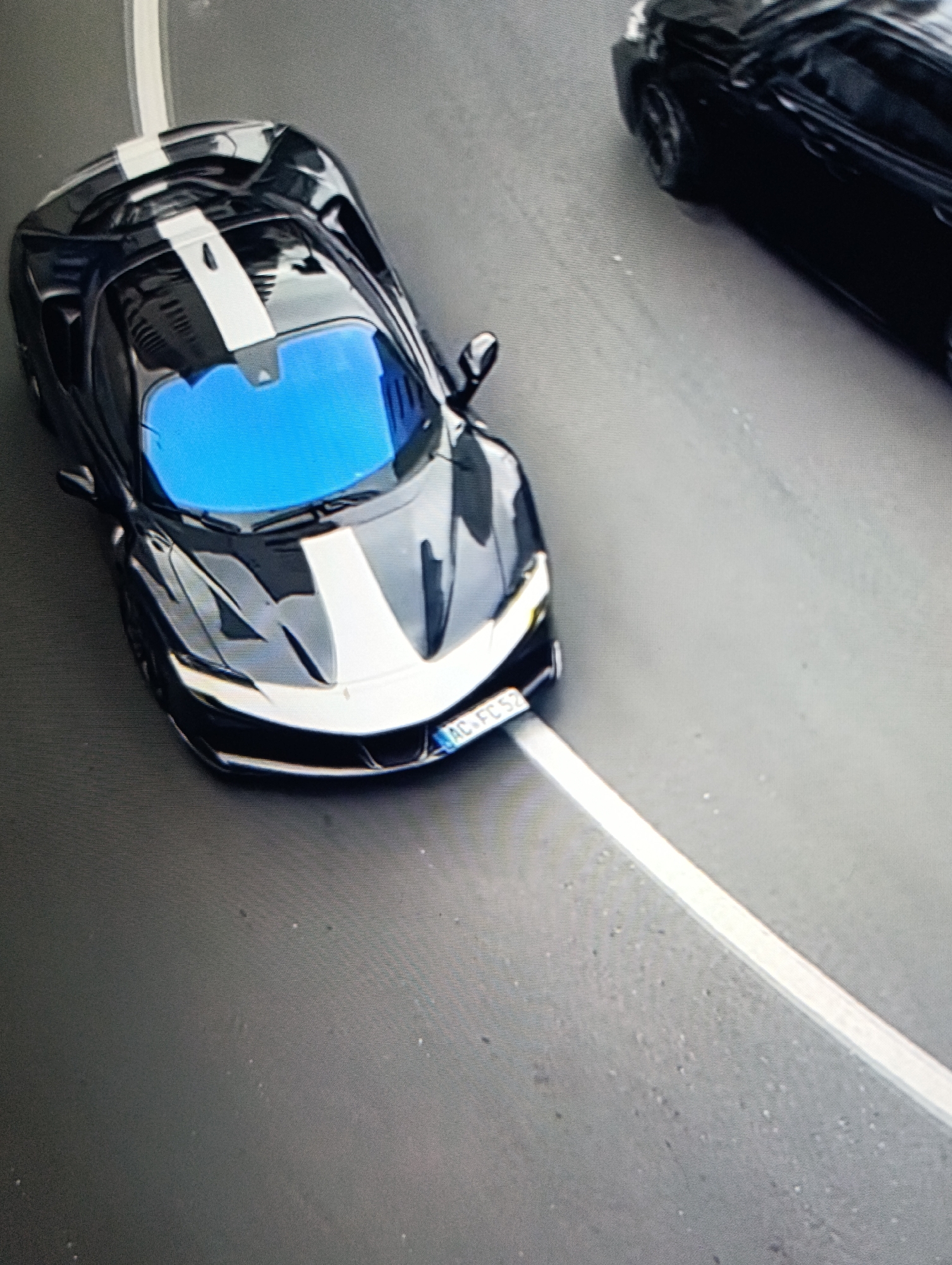 An aerial shot shows a black Ferrari 296 GTB with white racing stripes parked near a black SUV on a paved road in an urban area. The Ferrari's windshield reflects a bright blue sky. The road is marked with a white lane divider. The city and specific location are unidentified.