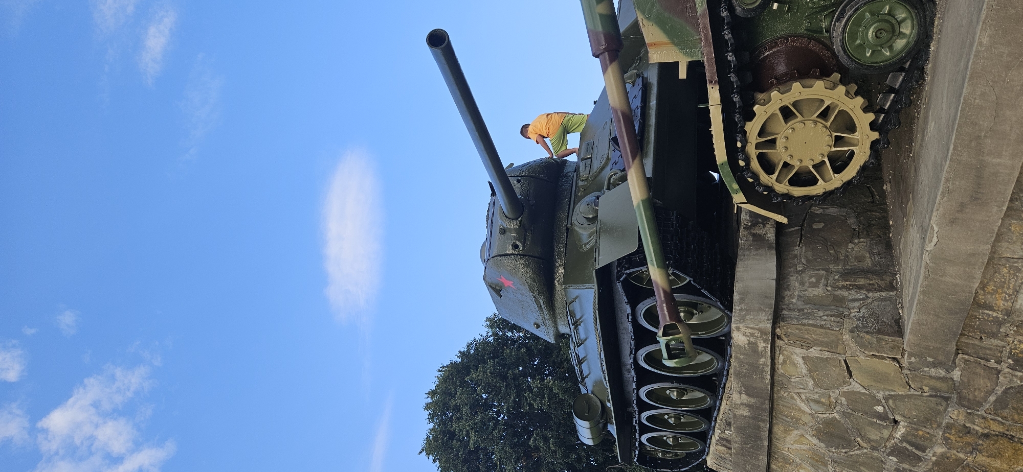 A low-angle, full-shot photograph captures two Soviet-era tanks on display, possibly as a memorial, against a bright blue sky with scattered white clouds. The tank in the foreground is partially obscured by the tank behind it. The dominant tank, a T-34, is painted dark green and has a prominent red star emblem on its turret. A young boy wearing an orange t-shirt and green shorts is perched on the turret of this tank, looking downwards. The tanks are positioned on a stepped, stone-like platform. The tank in the background exhibits a camouflage pattern of green, brown, and beige. The overall scene suggests an outdoor public space, likely in Svidník, Slovakia, known for its significant Soviet military history and monuments. The lighting indicates daytime, likely late morning or early afternoon.