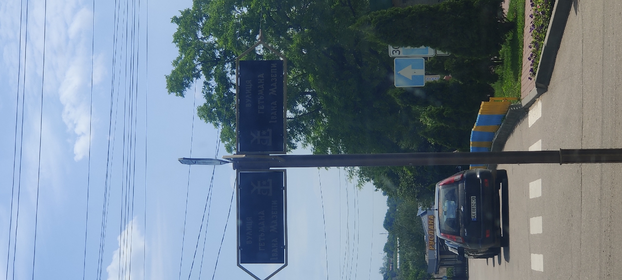 The image shows a street sign in the city of Kolomak, Ukraine, indicating directions to "ВУЛИЦЯ ГЕТЬМАНА ІВАНА МАЗЕПИ" (Hetman Ivan Mazepa Street). The sign is mounted on a pole with two black directional plates. The plate on the left points left, and the plate on the right points right. Both plates have the same text and a stylized symbol that appears to be a Cossack mace.
In the foreground, a black Chevrolet Aveo is parked, with its license plate visible as "AX 8852 OB". Behind the car, a colorful blue and yellow fence lines the side of the road. Further in the background, there are trees and buildings, including a sign with "РУСЛАНА" visible on a building. Traffic signs, including a directional arrow and a speed limit sign (30), are also present. The sky is blue with scattered clouds, and power lines are visible crossing the top of the frame. The scene appears to be on a bright, sunny day.