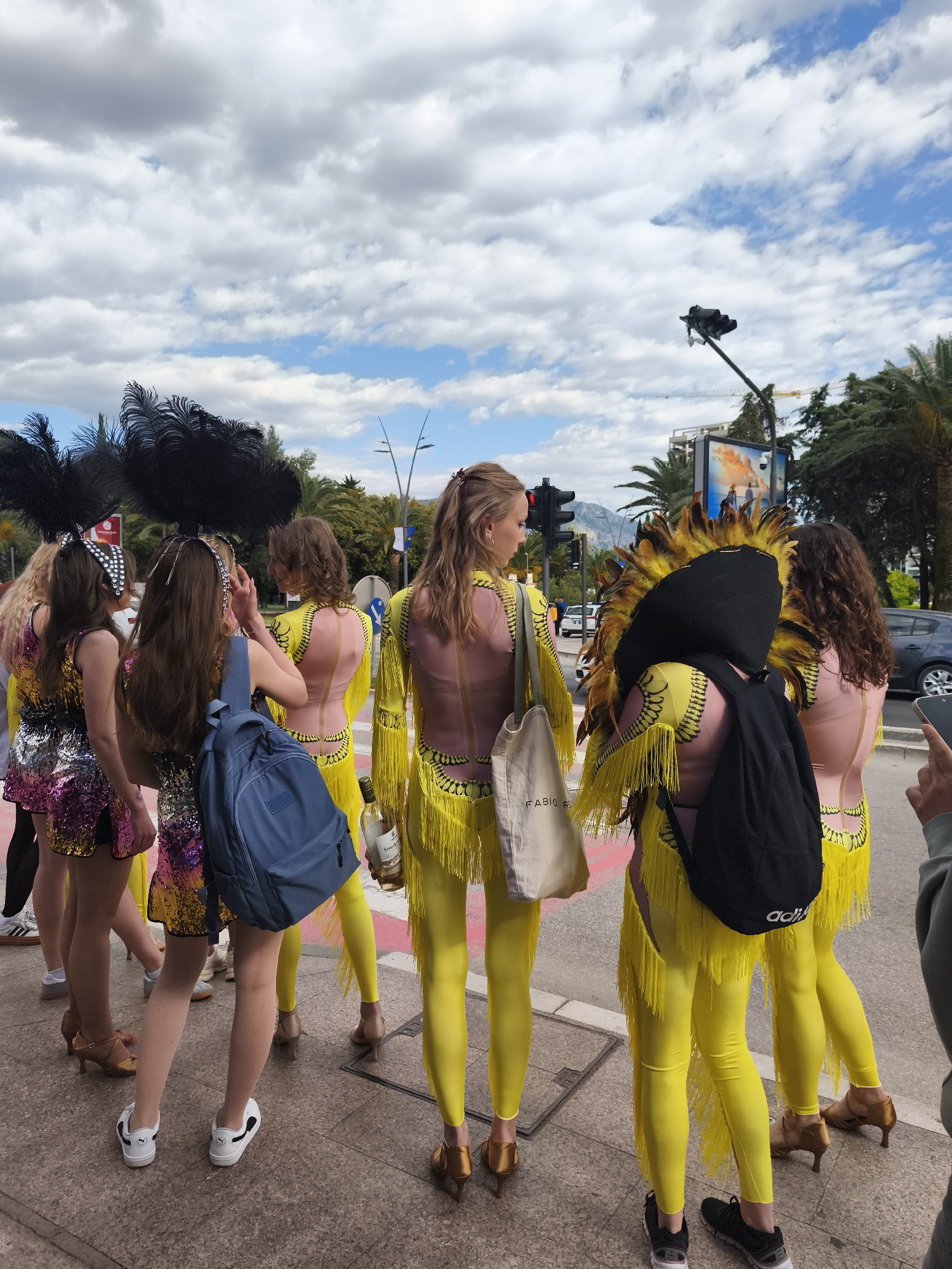 A street scene depicts a group of individuals, primarily women, dressed in colorful carnival-style costumes. The costumes vary but generally consist of fringed, brightly-colored outfits, some with feather headdresses. The individuals appear to be standing on a sidewalk or pedestrian area, with some holding bags or personal items. In the background, a city street is visible, with traffic lights, palm trees, and an advertising billboard in view. The sky is cloudy with patches of blue. The location is the city of Unknown, Unknown.