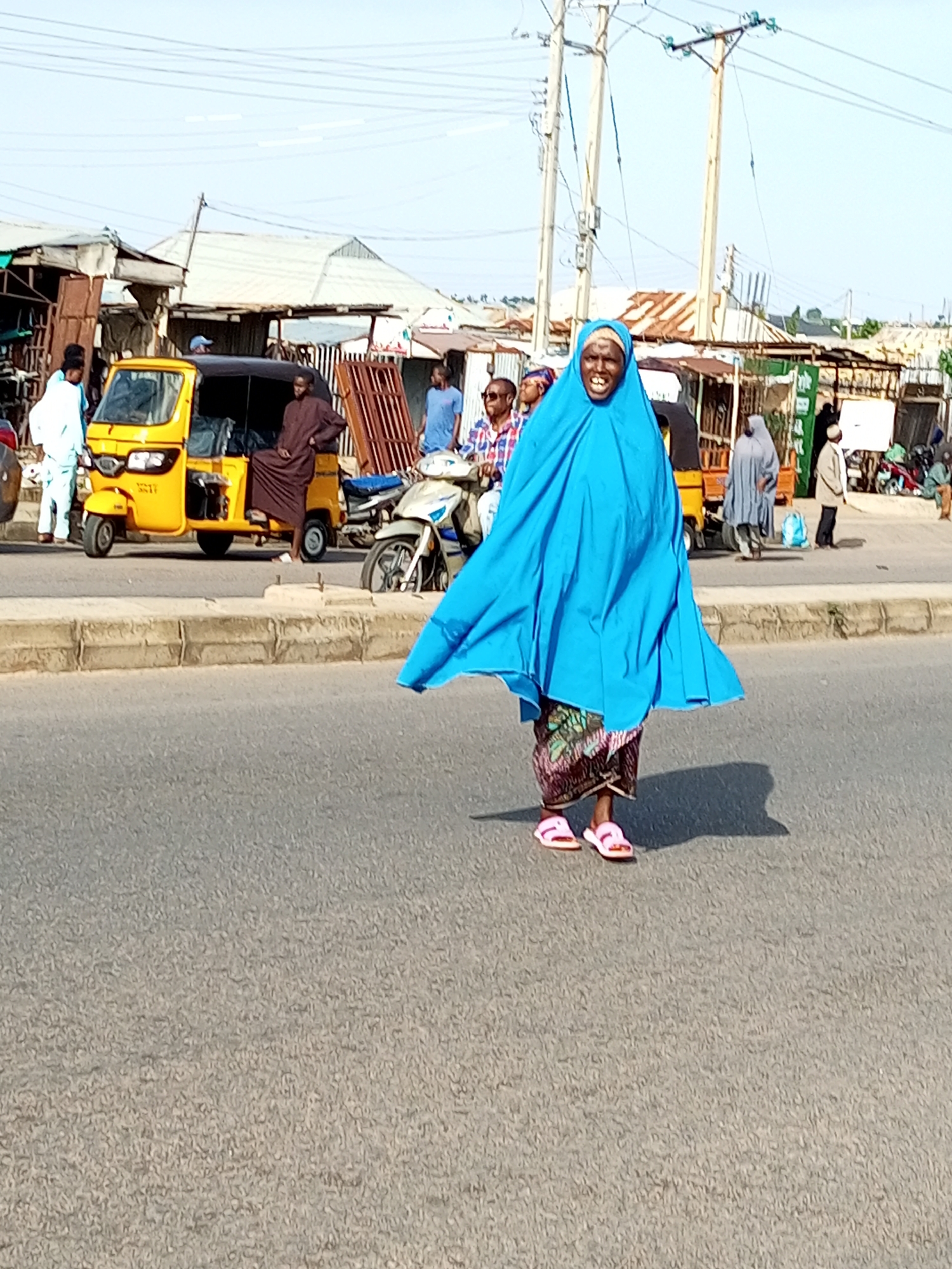 In the city of Tumfure, Nigeria, a woman wearing a vibrant blue hijab and a patterned skirt walks across a paved road. She is adorned with pink sandals. Her posture and facial expression suggest she is walking towards the viewer. Behind her, a yellow autorickshaw is parked on the side of the road, with a person wearing a brown garment seated on it. Further in the background, various market stalls and buildings are visible, alongside other individuals and a motorcycle. The scene is illuminated by bright daylight, casting shadows on the road surface. The environment appears to be a busy street, likely in a commercial area.