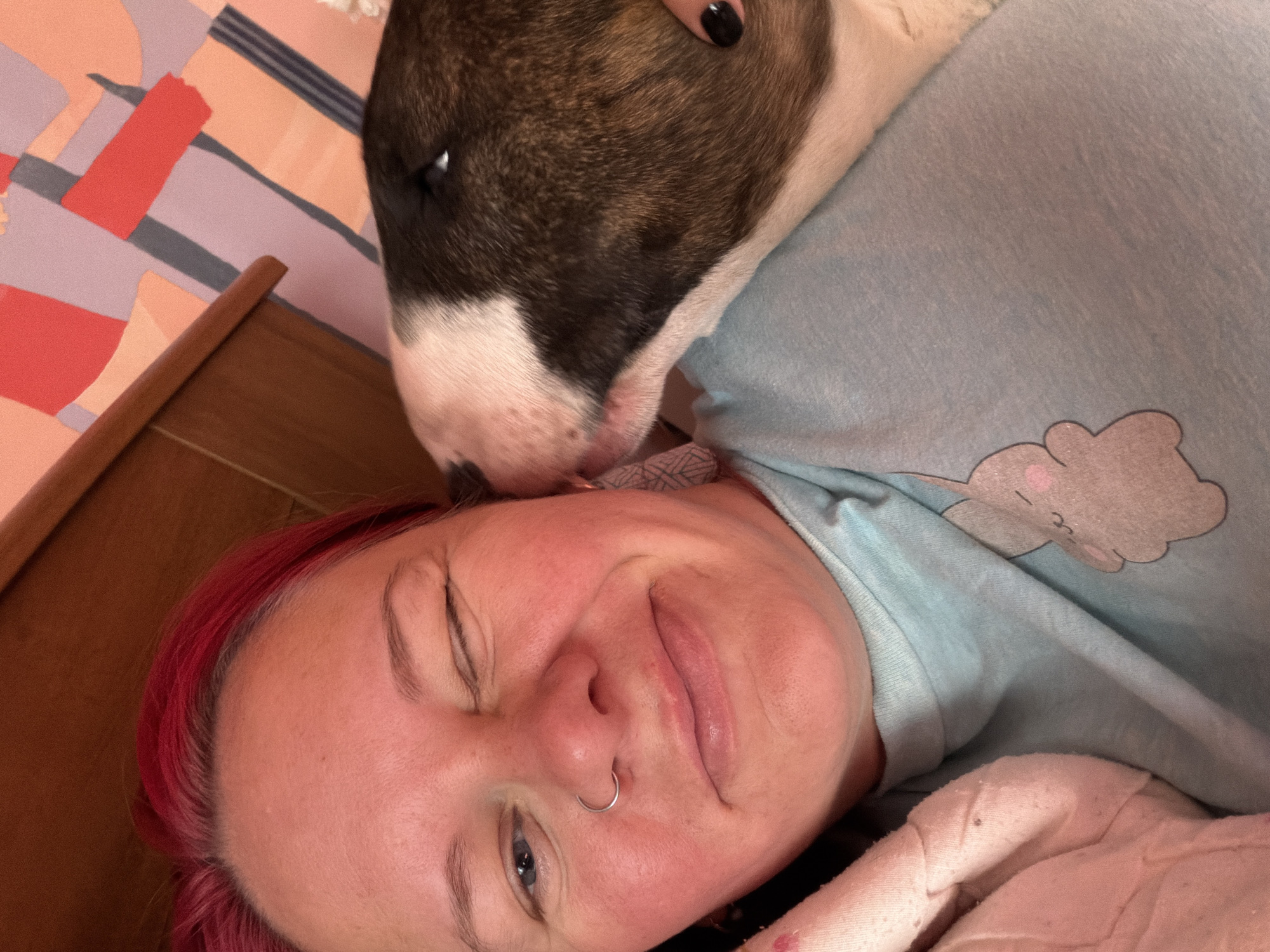 A close-up, high-angle selfie shows a woman with bright red hair winking as a bull terrier nuzzles her cheek. The woman is wearing a light blue t-shirt with a cartoon bunny printed on it and a silver nose ring. She is nestled under a pink blanket. The background includes a wooden headboard and a wall with abstract geometric patterns in shades of pink, orange, and blue. The location is not specified.
