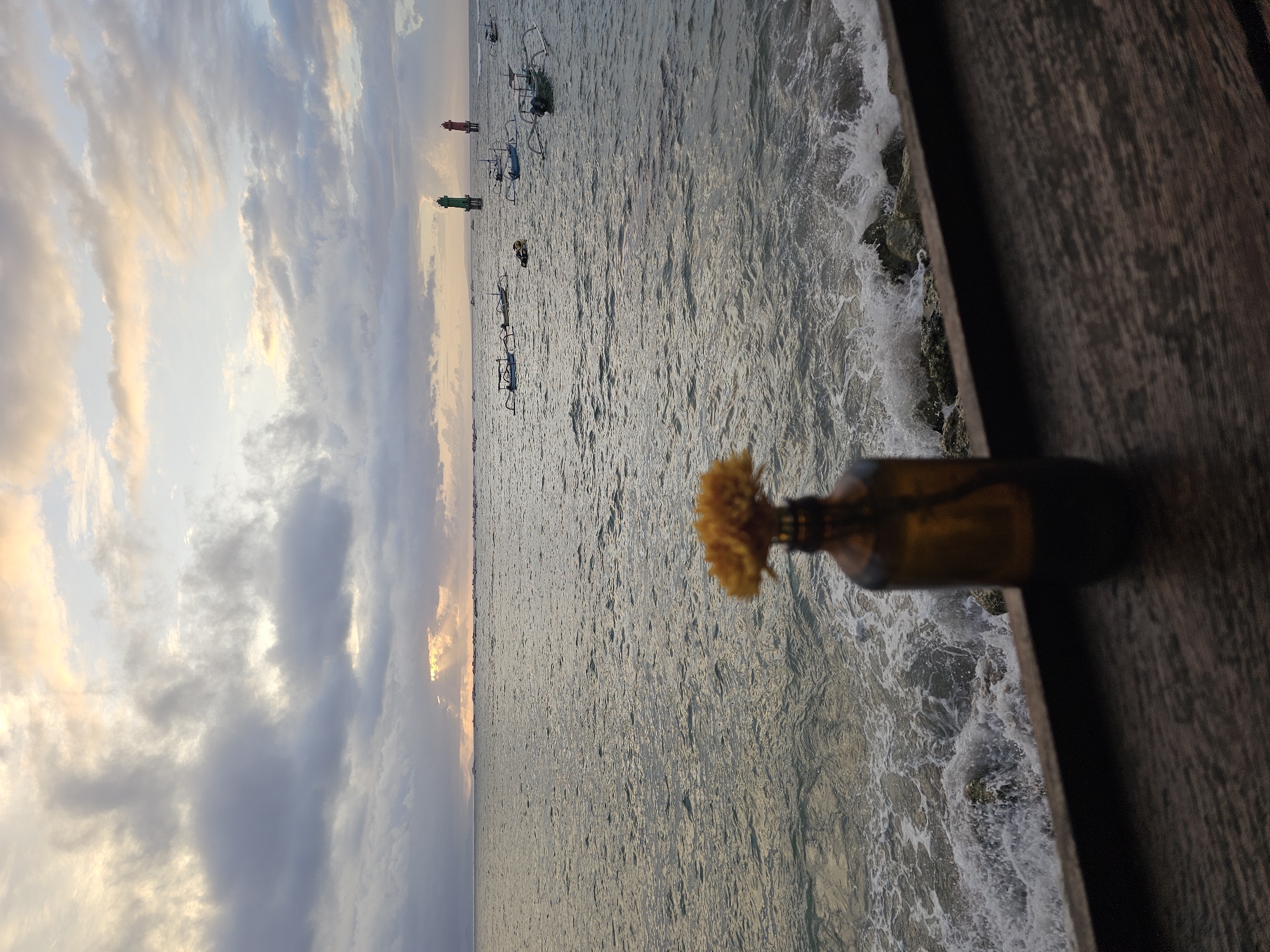 The image depicts a coastal scene in Denpasar, Indonesia. The sky is partially cloudy with hues of orange, suggesting a sunrise or sunset. The sea is visible, with waves crashing against rocks near the wooden edge of what appears to be a deck or pier. Several boats are anchored in the distance. In the foreground, a small brown bottle with a yellow flower protruding from its opening is positioned on the wooden surface.