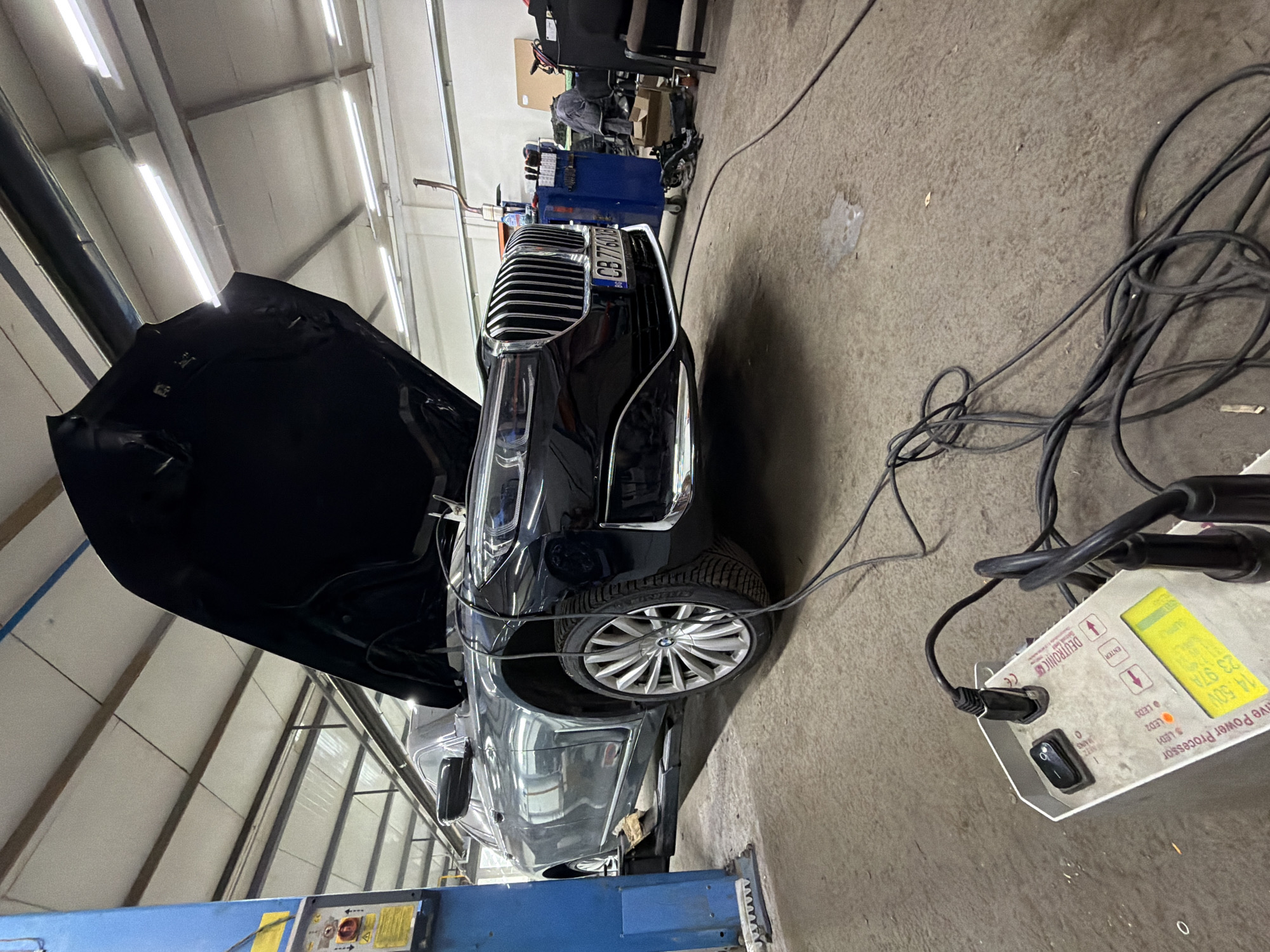 The image depicts a car repair shop in Burgas, Bulgaria, with a black BMW 7 Series sedan undergoing maintenance. The car is raised on a lift, and its hood is open, revealing the engine compartment. A diagnostic device, identified as a "Deutronic Live Power Processor," is connected to the vehicle, with cables running across the concrete floor. The device displays readouts, including "14.00V," "25.97A," and "TEST." In the background, there are various tools and equipment, including a blue toolbox and car parts. The environment appears to be a typical workshop setting, with industrial lighting and concrete flooring. No individuals are clearly visible in the frame.