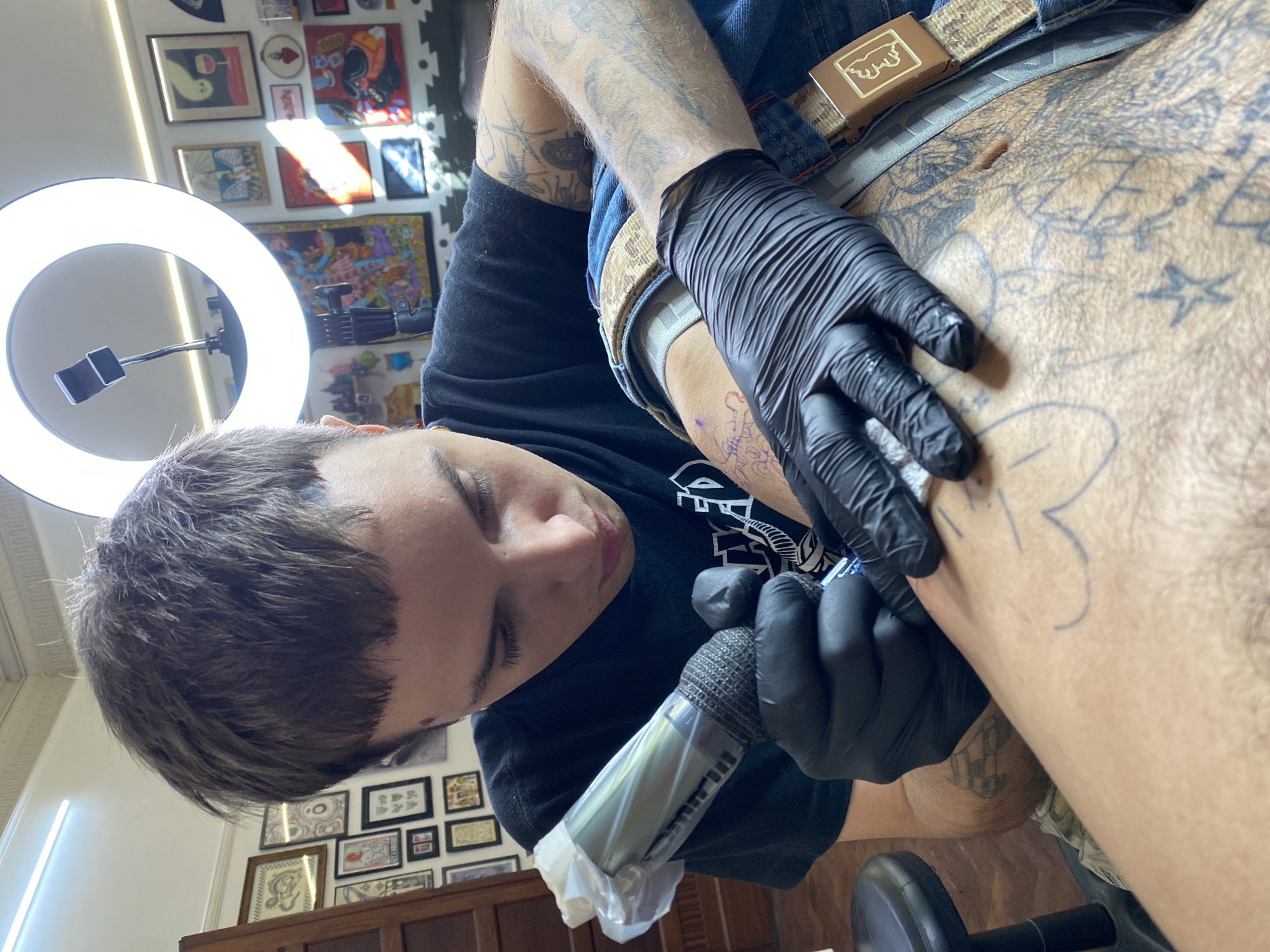 The photograph captures a tattoo artist in the process of tattooing a client's torso. The tattoo artist, a young person with short dark hair, is wearing a black t-shirt and black gloves. They are holding a tattoo machine, which is wrapped in a protective plastic cover. The client, who is wearing blue jeans and a belt with a brown buckle, has tattoos on their torso. The existing tattoos appear to be primarily black ink designs. The artist is actively tattooing the client's skin, adding to the existing body art.
The scene is illuminated by a ring light, creating a bright, focused light on the area being tattooed. The background reveals a wall adorned with framed artwork, suggesting the interior of a tattoo studio. There is a piece of dark wooden furniture visible in the background. A dumbbell is present near the bottom of the frame.
Without additional visual cues, it's impossible to definitively identify the location as Buenos Aires, Argentina. However, the presence of the Spanish language may be suggested by some of the art on the wall.