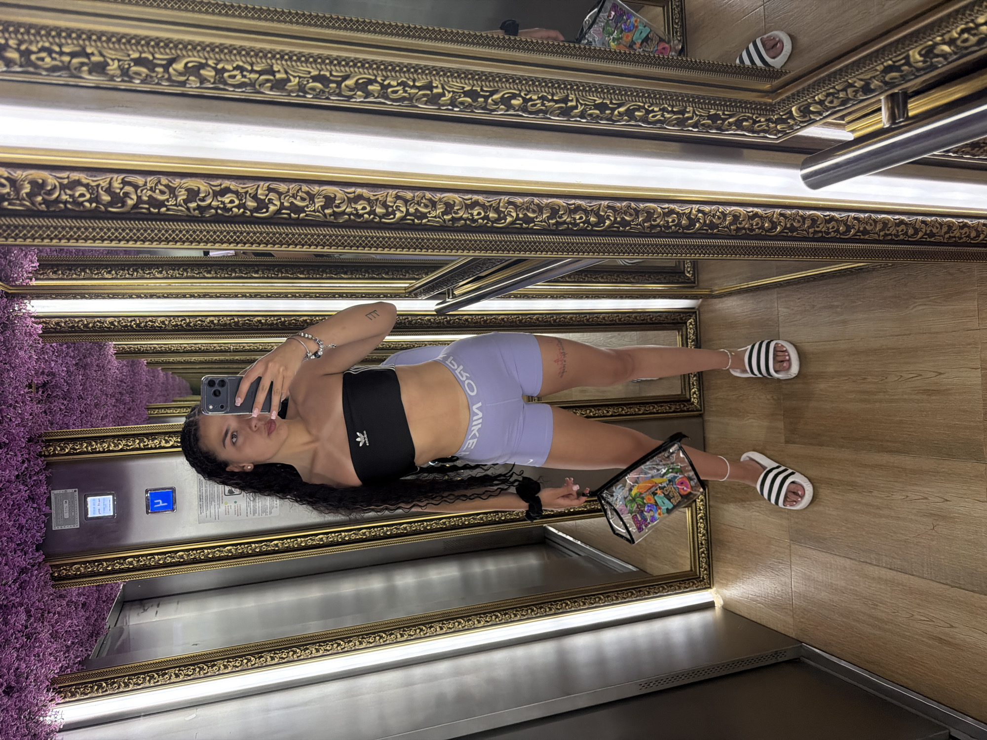 A young woman is taking a selfie in an elevator with multiple ornate mirrors. She is wearing a black Adidas strapless bra top and light purple Nike athletic shorts. She has long, dark curly hair and is holding a clear clutch bag with colorful stickers. She is wearing white Adidas slides. The elevator has a stainless steel door and a wooden floor. The ceiling is decorated with a dense, artificial purple floral arrangement. The mirrors reflect the woman, the elevator interior, and the floral ceiling. The elevator panel displays a blue illuminated "P" symbol.