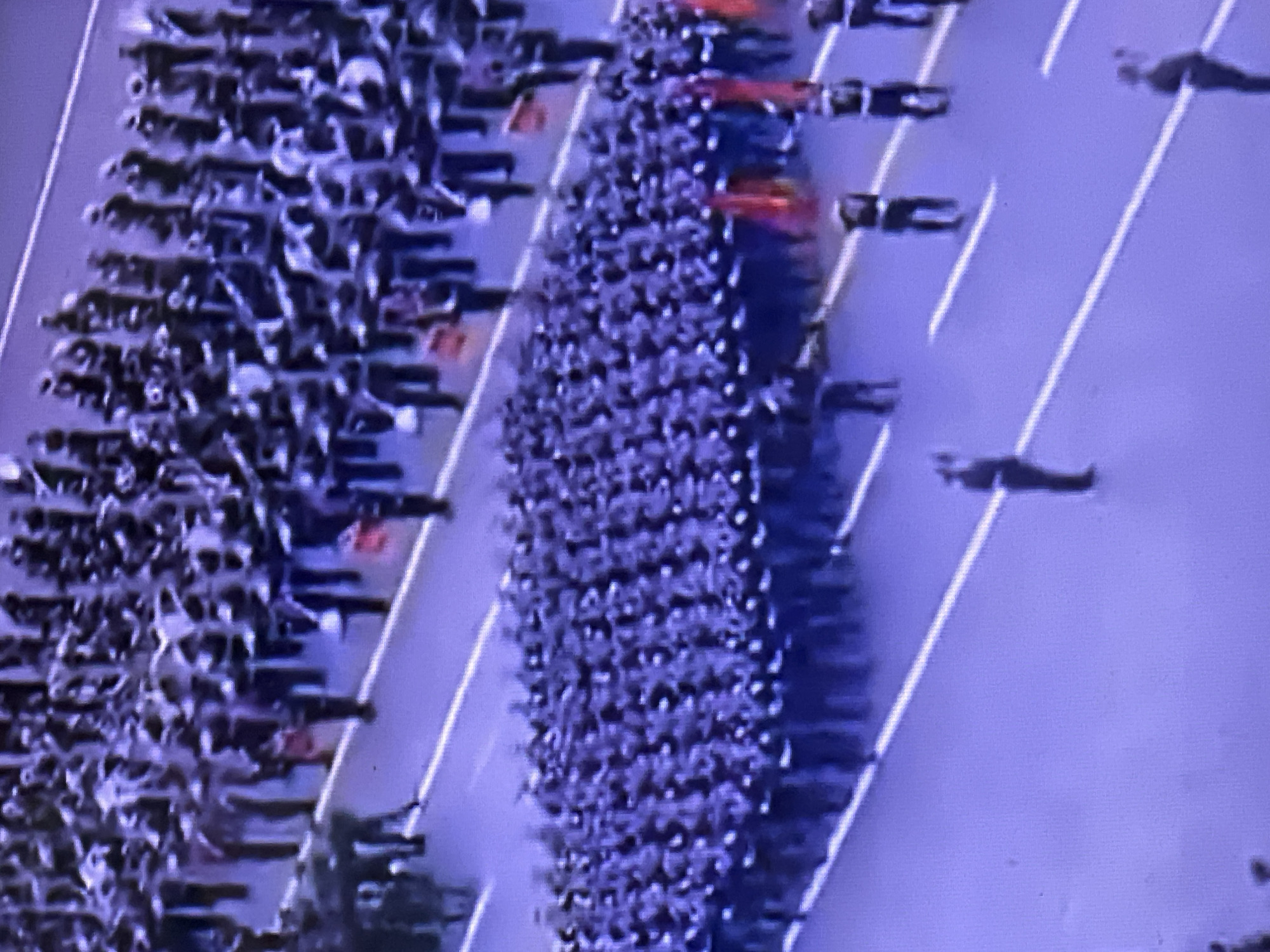 An aerial view shows a large military parade in Chaikovsky, Russia. Columns of soldiers march in formation on a wide street delineated by white lines. Some soldiers carry rifles. There are cars accompanying the march.