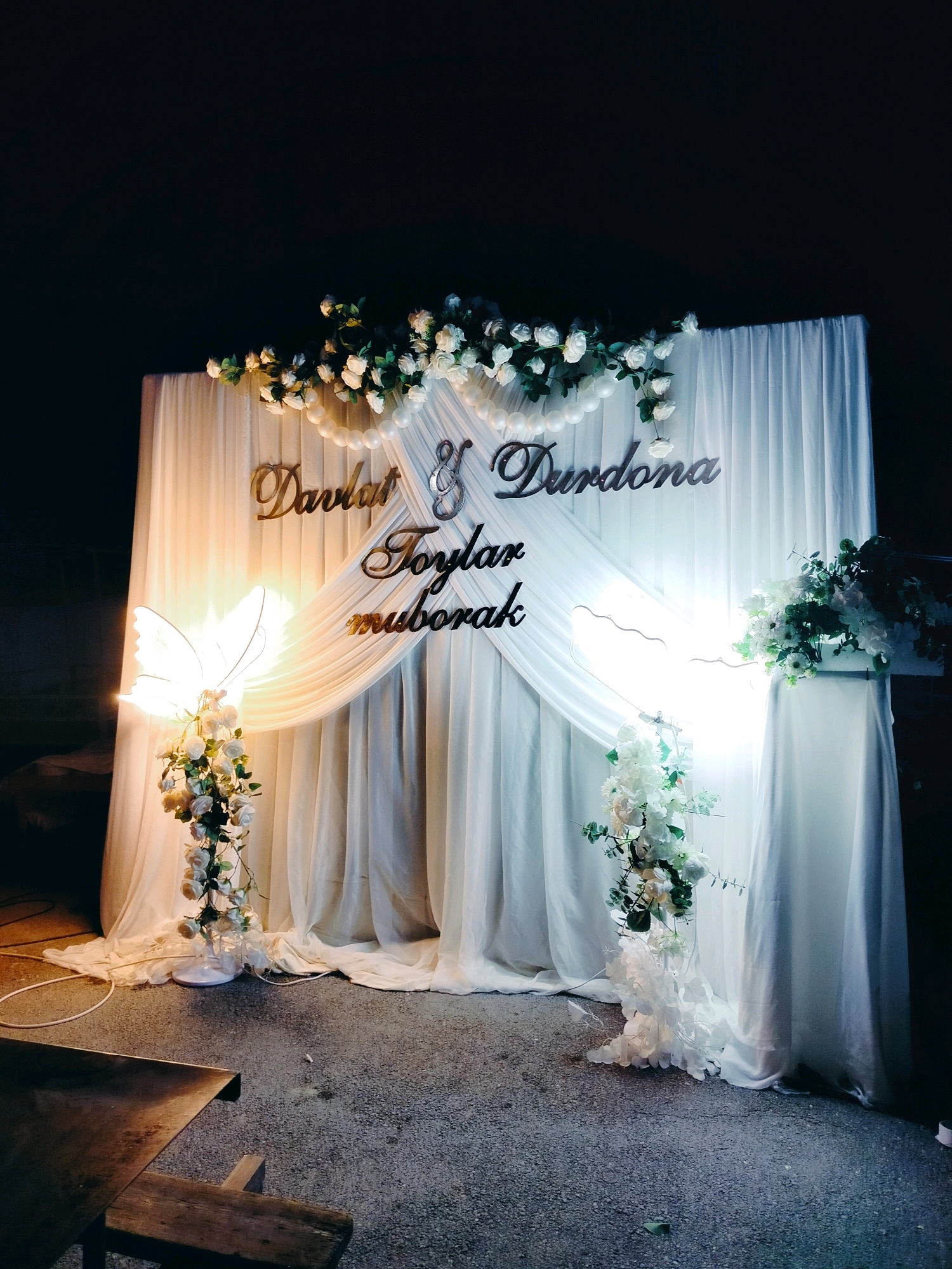 The image depicts an outdoor nighttime wedding backdrop in the city of Kölkent, Kazakhstan. The backdrop consists of draped white fabric, adorned with white roses and pearl strings. Prominently displayed on the fabric are metallic golden letters forming the names "Davlat & Durdona" and the phrase "toylar muborak," which translates to "congratulations on your wedding." To the left, a butterfly-shaped lamp illuminates a cascading arrangement of white roses. On the right, another floral arrangement, also featuring white flowers, is placed on a white pedestal. The ground is paved with asphalt, and in the foreground, a portion of a wooden table and bench is visible. The overall ambiance suggests a festive celebration.