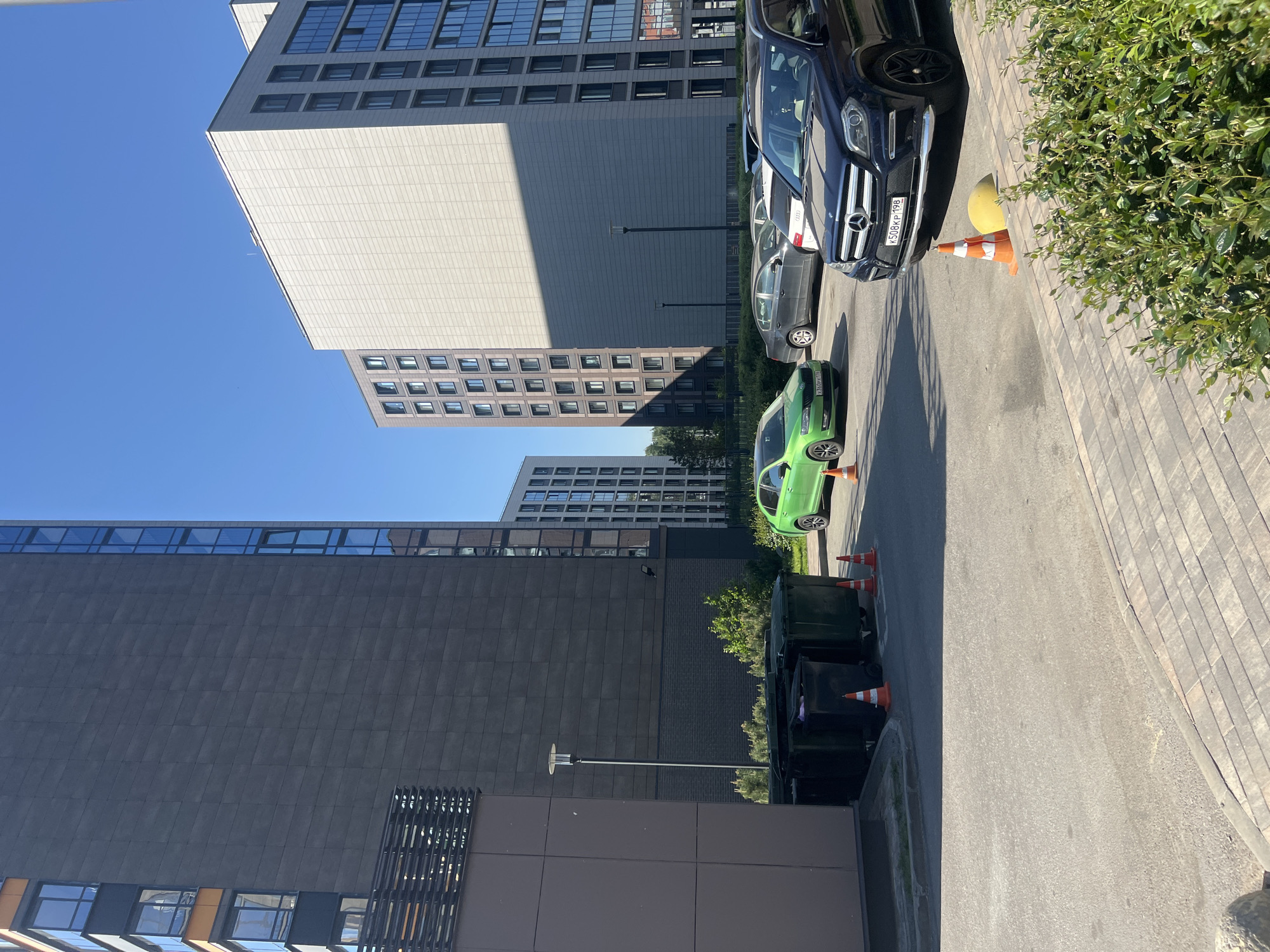 The image displays an urban street scene on a clear, sunny day in what appears to be a modern residential or office complex. In the background, several contemporary multi-story buildings dominate the skyline against a bright blue sky. The closest building on the left features a dark grey, paneled facade with many rectangular windows and a distinctive slatted architectural element. To its right, a lighter beige-grey building with a textured surface is bathed in sunlight, casting sharp shadows and revealing a grid pattern of windows.
The foreground consists of an asphalt road and a section of paved walkway or parking area made of light-colored paver blocks. Several cars are parked along the right side. A prominent dark blue/black Mercedes-Benz SUV is seen, with its Russian license plate "К508КР 198" clearly visible. Behind it, a bright green compact car, bearing the Russian license plate "У360РН 198," is partially obscured.
Multiple orange traffic cones are strategically placed near the parked vehicles and further down the street, close to a series of dark green trash bins. A large, spherical yellow object is partially visible next to the Mercedes. Green bushes and some small trees provide patches of foliage, breaking up the urban hardscape. Modern streetlights are also visible. The strong, direct sunlight creates distinct shadows, indicating a midday or early afternoon setting. The license plates confirm the location context of Saint Petersburg, Russia.