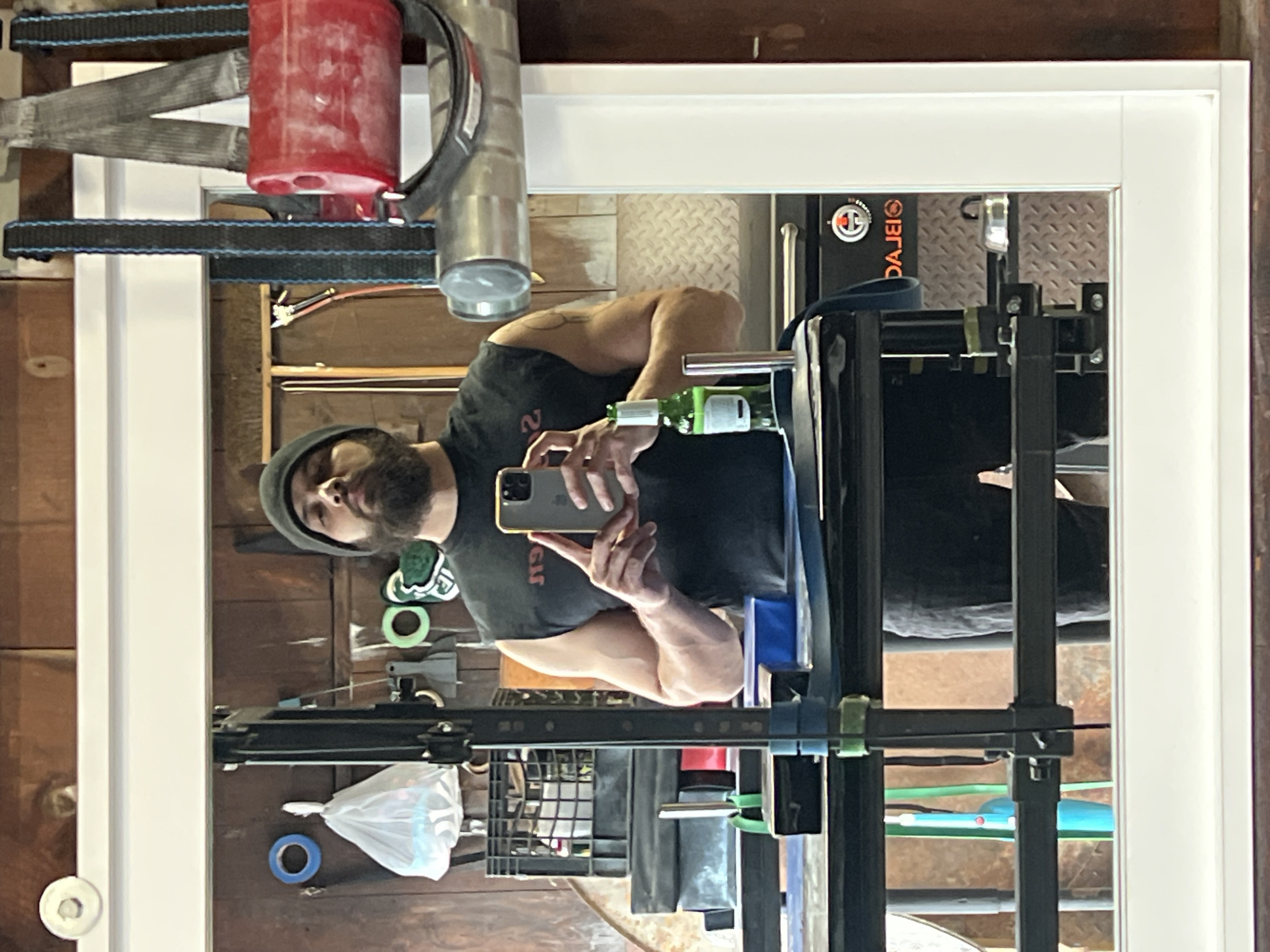 A man in a black shirt and gray beanie holds a smartphone, presumably taking a selfie, in front of a mirror. Reflected in the mirror is a garage or workshop setting. In the upper left corner, there is a red canister and metal equipment. Various tools, including tape and containers, are scattered on a wooden surface behind the man. A black metal rack is prominently displayed in front of the mirror. The location is presumed to be in the City of Newburgh, United States of America.
