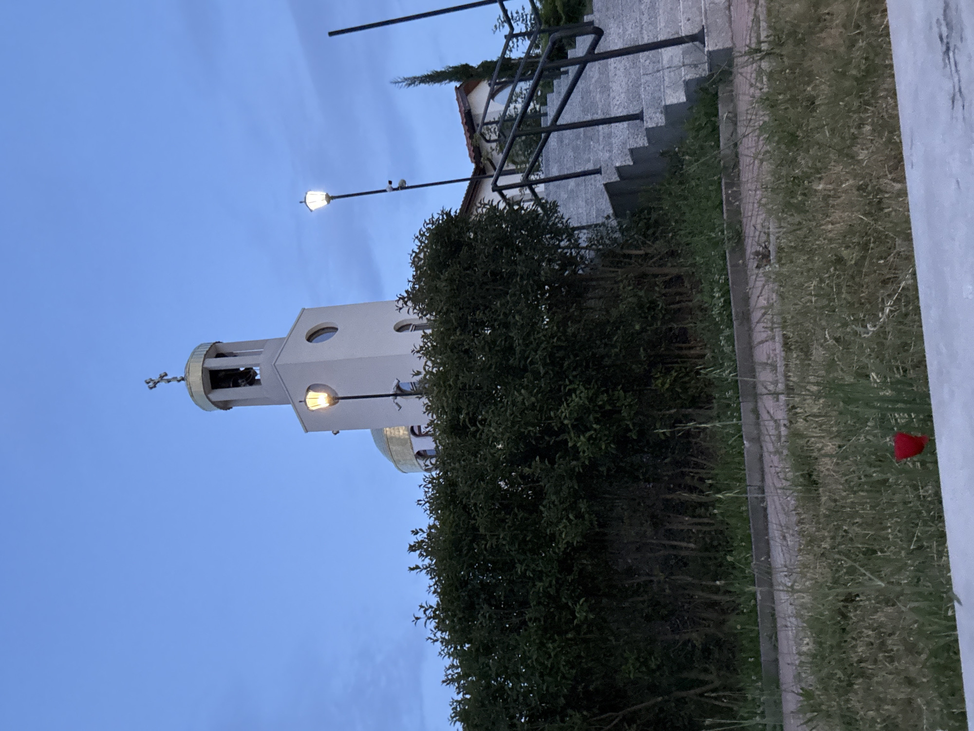 This image, captured in Sliven, Bulgaria, depicts an outdoor scene during dusk or early evening. The central feature is a white bell tower with a domed top, possibly golden, surmounted by an Orthodox cross. A bell is visible within the tower's belfry, and it features at least two circular windows.
In the foreground, a large, dense green bush partially obscures the lower part of the bell tower. To the right of the tower, a building with light-colored walls and a dark-tiled roof is visible, along with a set of concrete stairs equipped with a black metal railing. Two street lamps are present, both illuminated; one is positioned near the bell tower, and the other further right by the stairs.
The sky is a clear, deep blue, indicating twilight conditions. The ground level features grassy areas and smaller plants, with a paved edge or wall running along the right side of the frame. A single red flower, possibly a poppy, stands out in the grass near the bottom right. There is no visible activity or text in the image.