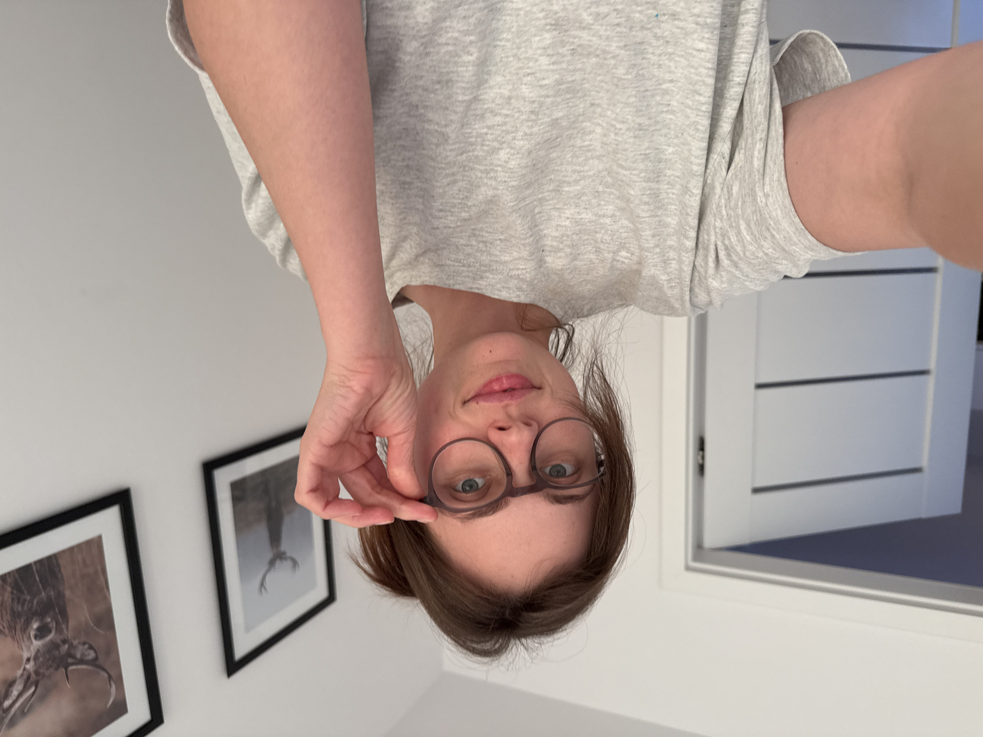 This is a portrait-oriented, slightly angled, selfie-style photograph of a young woman wearing glasses and a grey t-shirt. The image is upside down, so the woman's head is at the bottom of the frame and her feet (implied) would be at the top. She is looking directly at the camera with a neutral expression. Her hand is raised and she is touching the frame of her glasses with her index and thumb. Two framed pictures of deer hang on the wall behind her to the left. The room appears to be well-lit, suggesting it is daytime. The setting is likely a home or indoor space. There is no visible text in the image. The location context provided is Szklarska Poręba, Poland.