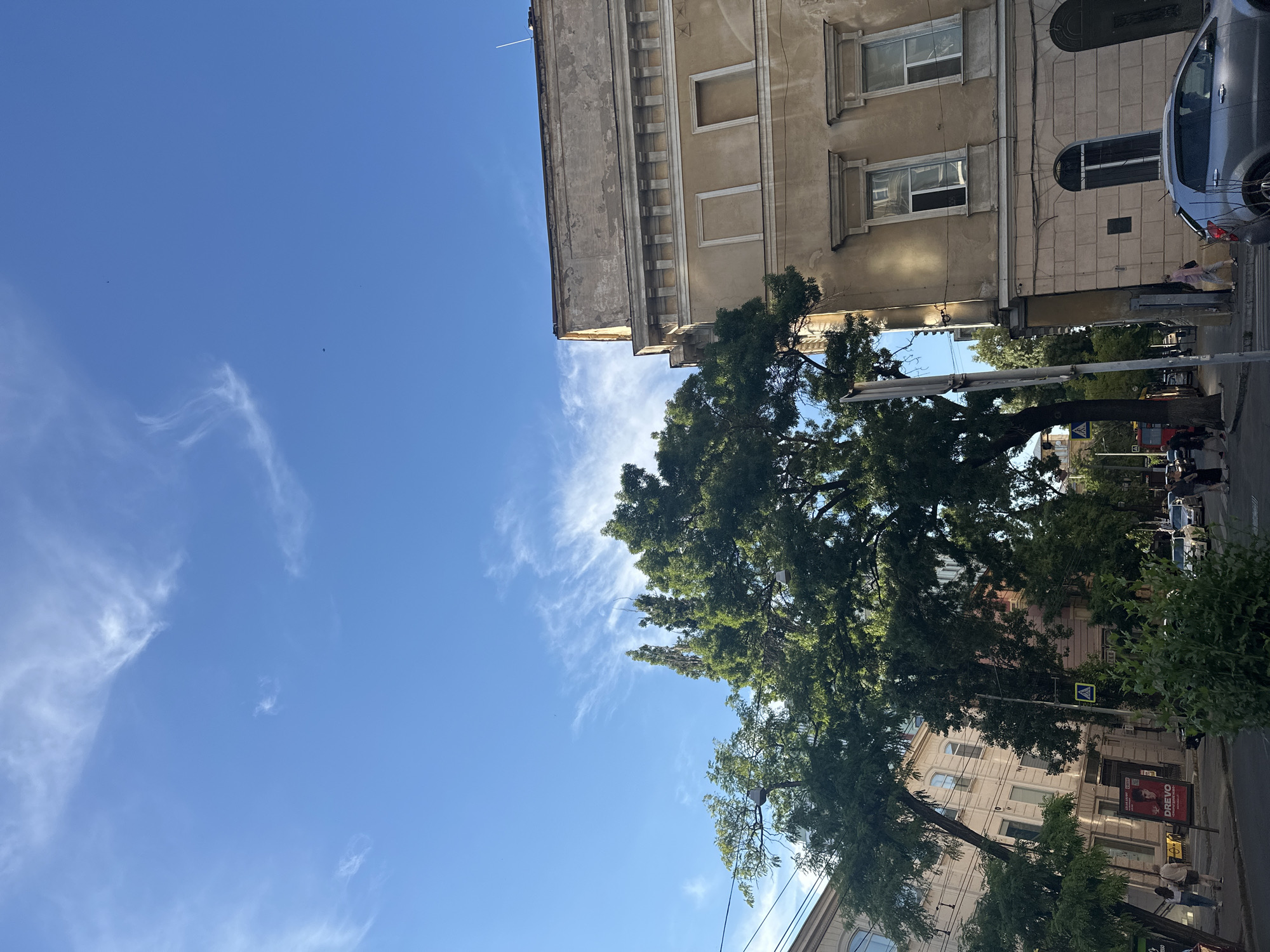 This image captures a street scene in Odesa, Ukraine, on a clear day with a bright blue sky adorned with wispy white clouds. The perspective is from a street level, looking up and diagonally towards the right. Prominently featured on the right is a multi-story building with a weathered, ochre-colored facade, characterized by classical architectural elements such as ornate window frames and a decorative cornice. The building appears to be an older structure, showing signs of age.
To the left, other buildings are visible, one with a light beige facade and arched windows, and further back, a building with a darker facade. Lush green trees are interspersed throughout the scene, with a large, mature tree dominating the mid-ground, its branches extending towards the buildings. Some of these trees are leaning, suggesting they are quite old.
In the foreground, a street runs along the bottom of the frame. Cars are parked along the street, including a silver hatchback visible on the right. Pedestrians are present on the sidewalk and crossing the street. A street sign with a yellow yield symbol is visible on the left side of the street, indicating a crosswalk. A red bus is partially visible in the background on the left. Advertisements are displayed on the side of one of the buildings. The overall impression is of a vibrant urban environment with a blend of historic architecture and natural greenery.