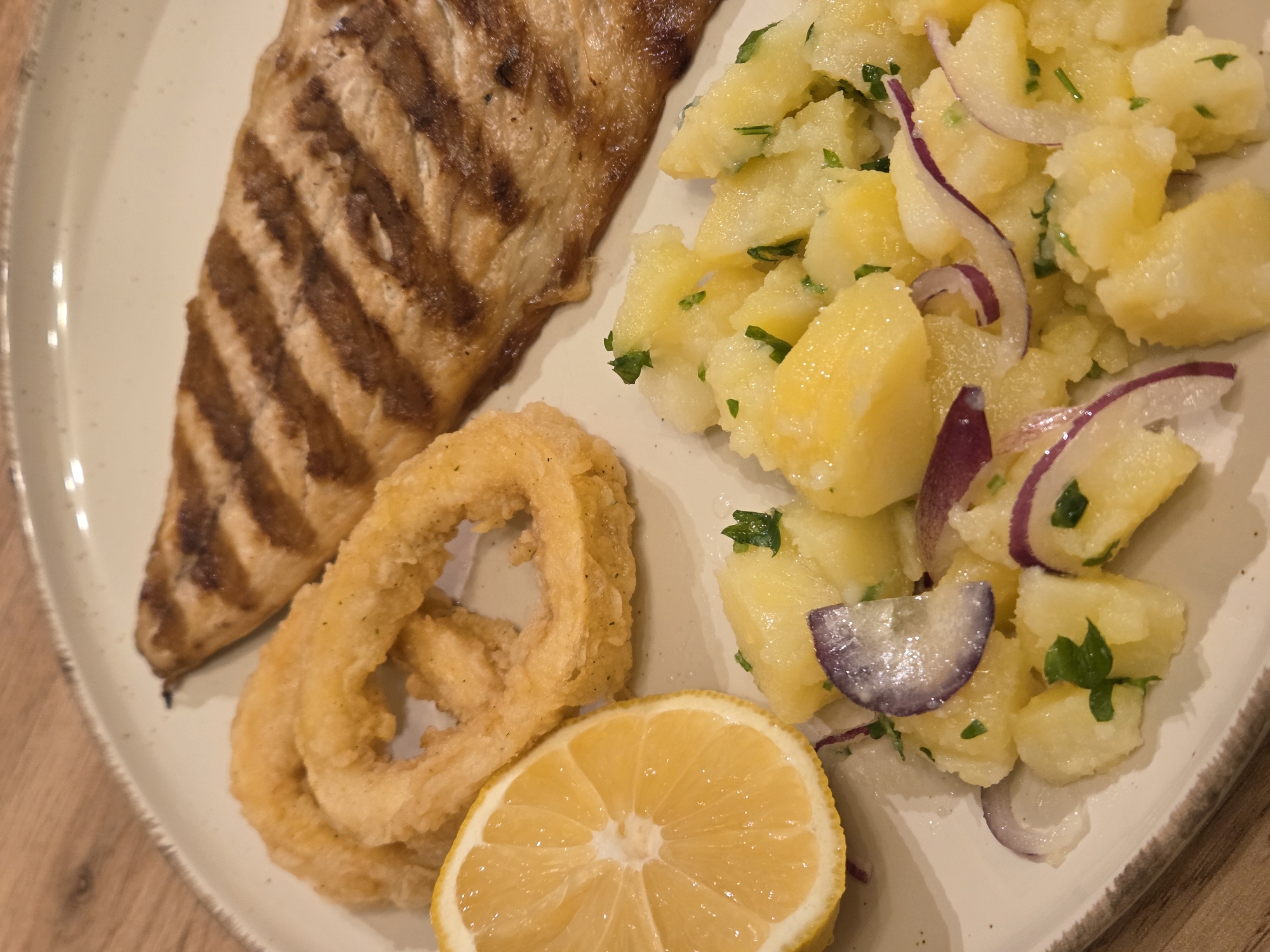 The image shows a plate of food, likely in a restaurant setting, though the precise location within Varna, Bulgaria cannot be determined. A grilled fish fillet with prominent grill marks is on the left. Next to it are two fried calamari rings and a lemon wedge. A potato salad with chopped herbs and thinly sliced red onions occupies the right side of the plate. The plate is light-colored with a textured rim, and the table underneath is a light-colored wood. No people are visible in the image.