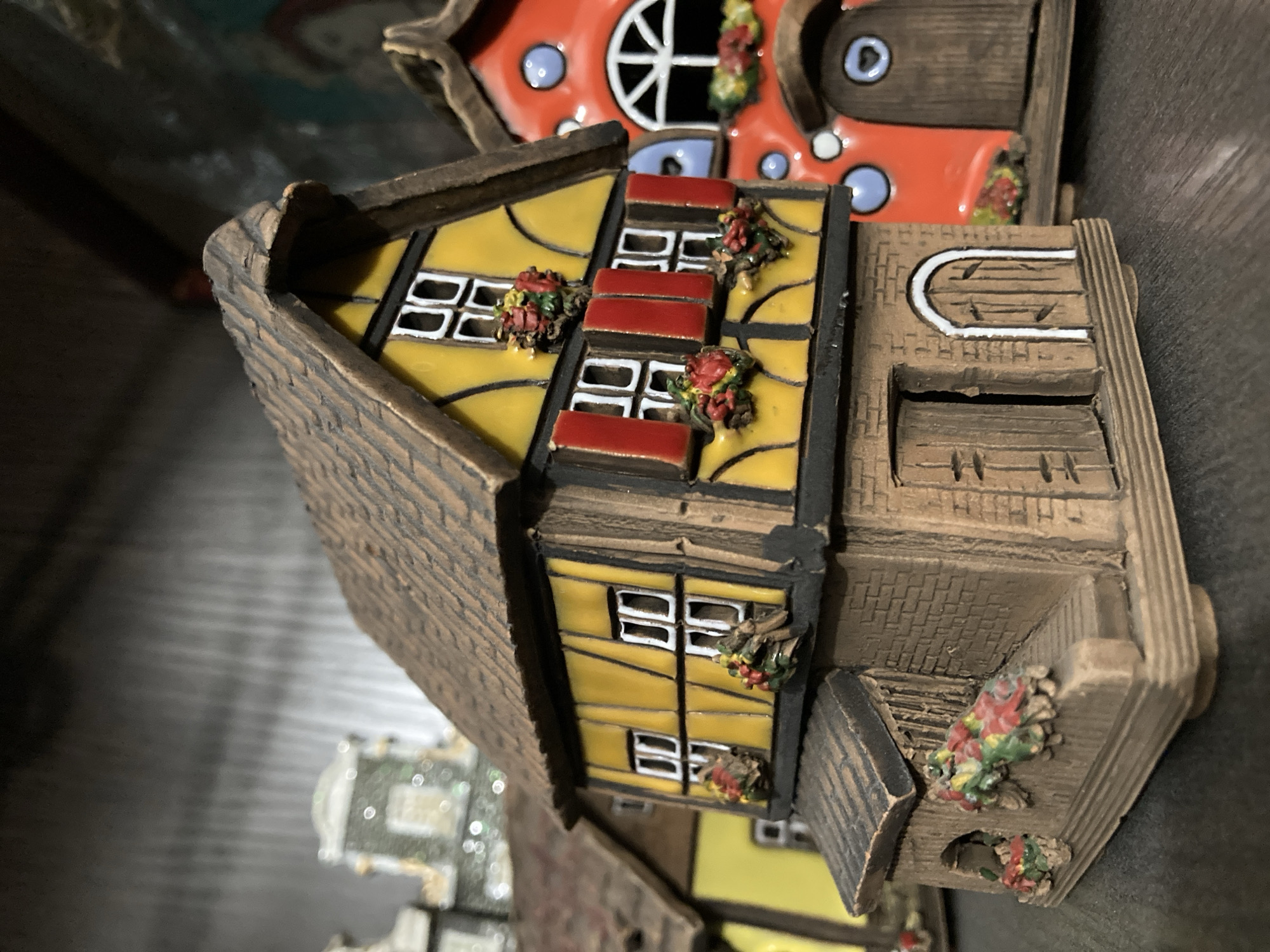 This is a close-up, eye-level, indoor shot of a display of miniature houses, possibly figurines. The main subject is a detailed, multi-story, half-timbered house with a yellow facade, dark brown beams, and a brown, textured roof. It has several windows with white panes and red shutters. Small, colorful flower decorations adorn the window sills and the lower portion of the house. To the right of the main house, a bright orange house with a rounded top and blue circular accents is partially visible. In the background, a blurred, light-colored building with a domed roof is also visible. The houses are placed on a dark, flat surface, likely a shelf, with a dark, out-of-focus background. The lighting is dim, highlighting the textures and colors of the miniature houses. There are no people or animals in the image. The scene is static and appears to be a still life arrangement.