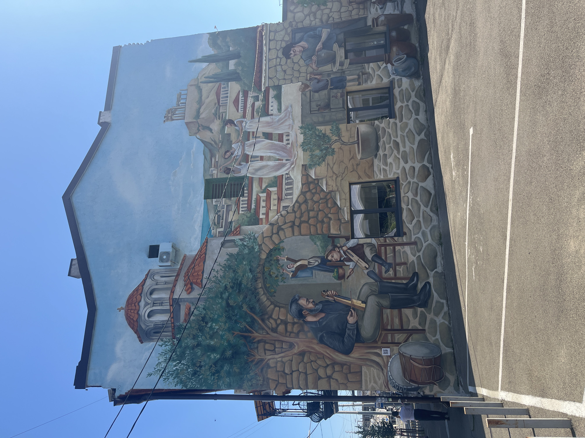 The image shows a large, colorful mural on the side of a building. The mural depicts a scene with elements of Greek architecture, including the Acropolis in the background, and several figures engaged in activities such as playing music, dancing, and pottery. A traditional Greek church with a red dome is also visible. The mural is painted on a light blue sky with some clouds.
The building appears to be located in a city, as evidenced by the power lines and a street visible in the foreground. A person in a white shirt and dark pants is walking on the sidewalk to the left of the mural. Parking lines are painted on the asphalt in front of the building.
This mural is located in the city of Krasnodar, Russia. Specifically, it is on the building at 89 Krasnaya Street. The mural was created in 2022 by artists from the "Art City" project and is intended to evoke a Mediterranean atmosphere.