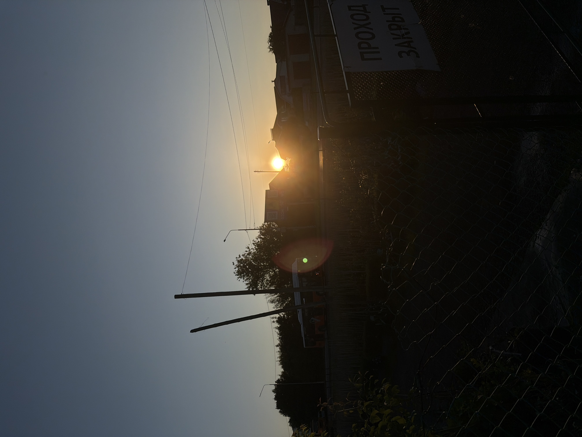 The image depicts a rural-urban transitional area in Новинки, Russia, at either sunrise or sunset, characterized by strong backlighting from a low sun. The bright, obscured solar disk is positioned above the horizon, slightly right of the frame's center, emanating significant lens flare with green, red, and orange concentric rings. The sky above is a clear, gradient blue, transitioning to warm yellow and orange hues near the horizon.
In the foreground, a dark, wire mesh fence (chain-link) runs diagonally from the lower-left to the center-right, casting deep shadows. To the far right, a solid dark metal gate post or barrier is visible, bearing a rectangular white sign with dark Cyrillic text. The visible portion of the text reads "ПРОХОД ЗАКРЫТ," which translates to "Passage Closed" or "Entry Closed." Dark, out-of-focus foliage occupies the extreme lower-left corner.
The midground features a road, mostly in shadow, traversed by a white and orange urban bus, partially silhouetted against the lighter sky. The number "15" is faintly visible on the bus's front, suggesting a route number. Two tall, slender, dark poles, possibly utility poles or temporary supports, lean slightly inward in the central-left area. Behind the road, a line of dark, dense trees creates a prominent silhouette against the horizon on the left.
On the right side of the midground, several low-rise buildings, likely residential houses or small commercial structures, are silhouetted, with some revealing peaked roofs. Overhead, power lines stretch horizontally across the frame. A faint "OZON" logo, indicative of an e-commerce platform, is discernible on a sign above one of the buildings on the mid-right. No individuals are explicitly visible in the scene.
