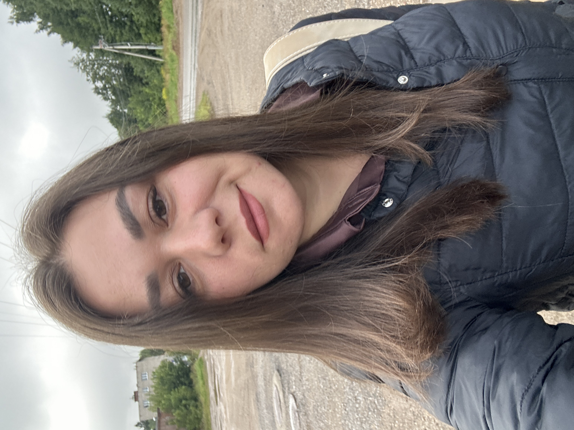 A young woman with long, dark brown hair is taking a selfie outdoors. She is wearing a dark blue puffer jacket and a maroon collared shirt. A light-colored strap, likely from a bag, is visible over her shoulder. The background shows a cloudy sky, a few trees, a power line pole, and a building. The ground is rough and appears to be a gravel or dirt road with some puddles. The scene is likely in Chaikovsky, Russia, given the natural and somewhat rural setting with a modest building in the distance.