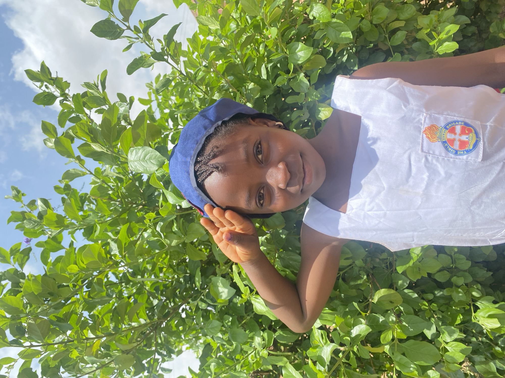A young black girl wearing a blue hat and a white shirt with a "Girls Brigade" patch stands in front of a lush green bush on a partly sunny day. The location is likely Turu, Nigeria.