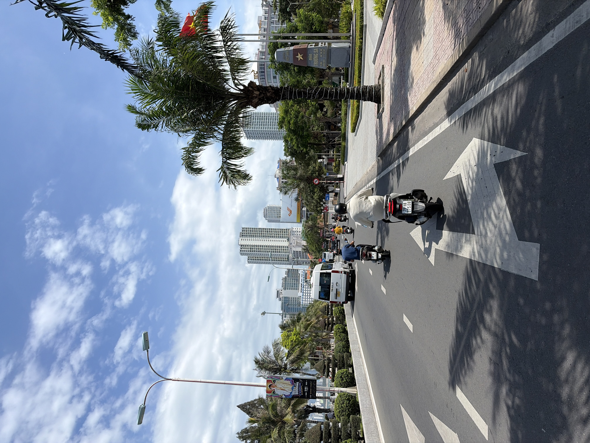 The image depicts a sunny day in Nha Trang Ward, Vietnam, featuring a street scene with several motorbikes and a white van driving on a multi-lane road. The road has white lane markings, including a prominent left-turn arrow. On the right side of the road, there is a sidewalk with patterned paving stones, a palm tree with its trunk wrapped in white material, and a monument with Vietnamese text and a star emblem. Behind the monument, a Vietnamese flag flies from a flagpole. A line of smaller trees and bushes separates the sidewalk from a line of modern, multi-story buildings in the background. On the left side of the road, there are more palm trees, a street lamp with two fixtures, and a billboard advertising an event. The sky is blue with scattered white clouds. The overall impression is one of a vibrant urban environment with lush greenery and clear weather.