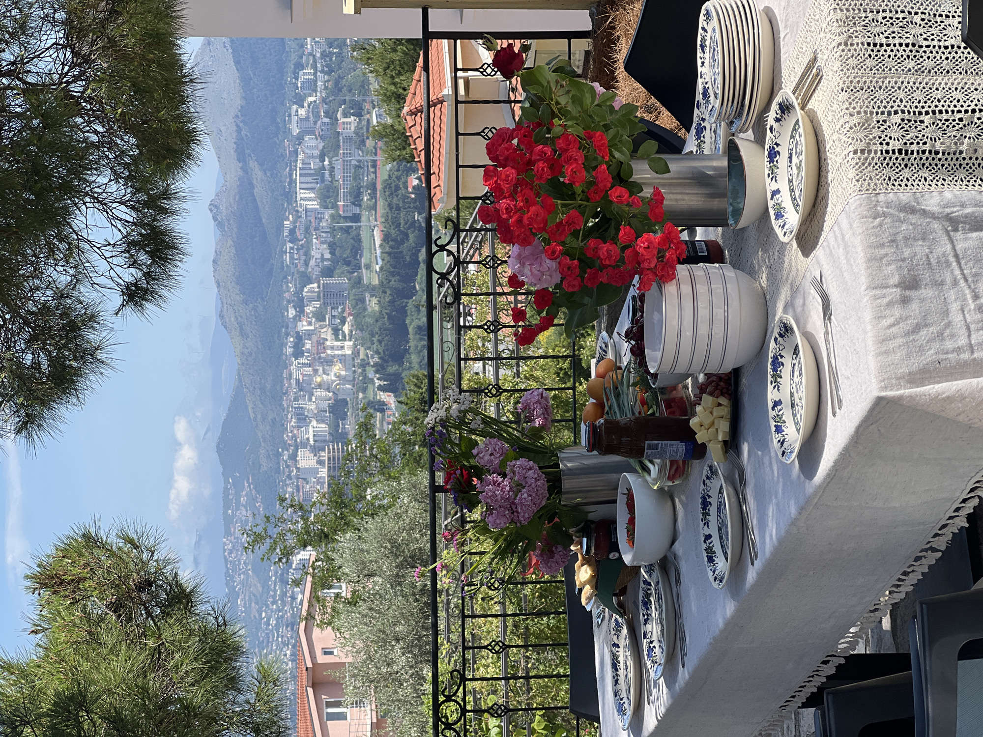 The image captures an outdoor dining scene with a view of the city of Šušanj, Montenegro, and the surrounding mountains. The setting is a balcony or patio, indicated by the wrought iron railing and surrounding vegetation. A long table dominates the foreground, draped with a white tablecloth, and set for a meal. Stacks of blue and white patterned plates, cutlery, bowls, and serving dishes are arranged along the table. Decorative flower arrangements in silver containers are positioned near the table’s edge, adding color and visual interest. In the background, the city of Šušanj is visible, nestled along the coast, with numerous buildings and a complex urban layout. Mountains rise in the distance, under a blue sky. Trees frame the left side of the image. No people are directly visible.