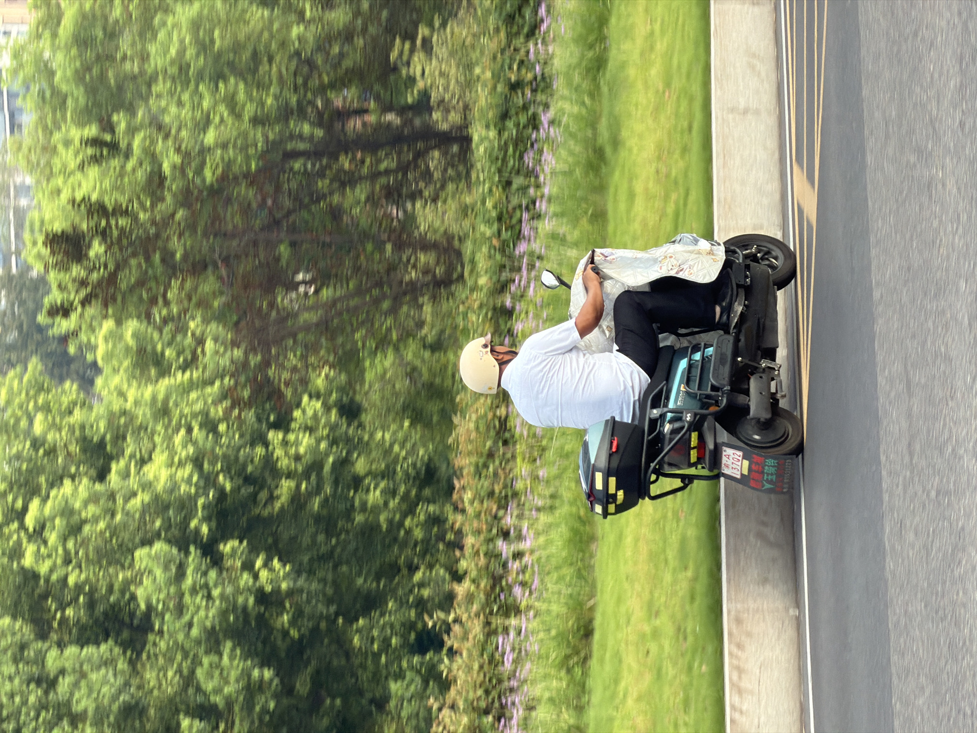 In the city of Jiangjin, China, on a paved road with yellow lane markers, a person wearing a white shirt, black pants, and a light-colored helmet rides a turquoise electric scooter. The scooter has a black storage box on the back with yellow reflective stickers. The rider is partially covered by a patterned cloth. The background features a grassy embankment with scattered purple flowers and a dense line of green trees. Buildings are visible in the far distance behind the trees. The scene appears to be captured from a moving vehicle, indicated by the motion blur in the background.